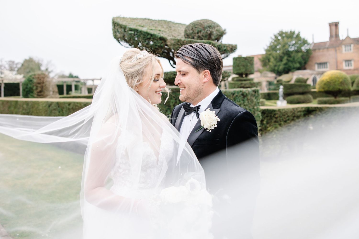 A bride in a flowing veil and groom in black tuxedo share an intimate moment in front of a garden setting.