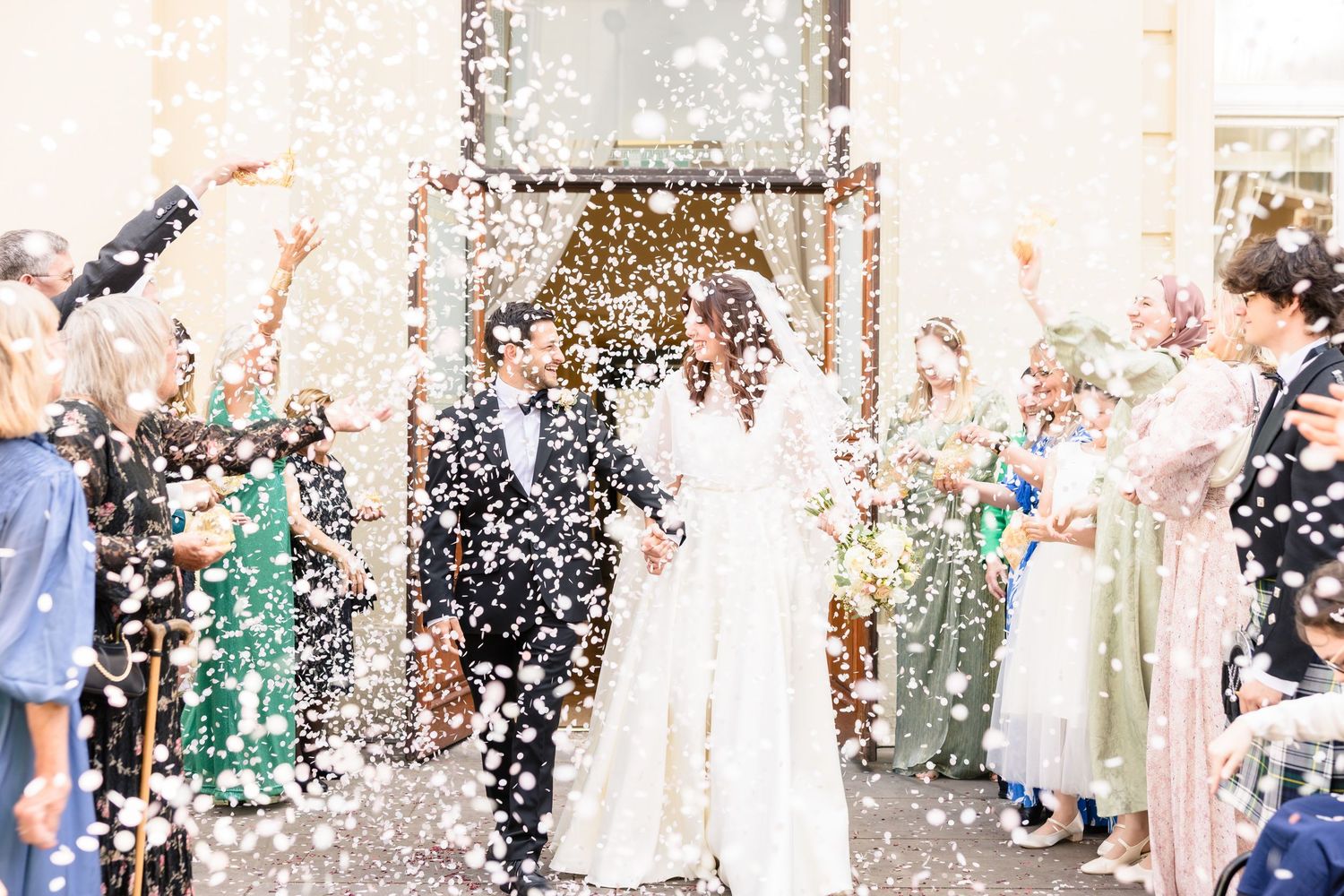 Wedding party poses for photos while guests throw white confetti in celebration at an outdoor venue.