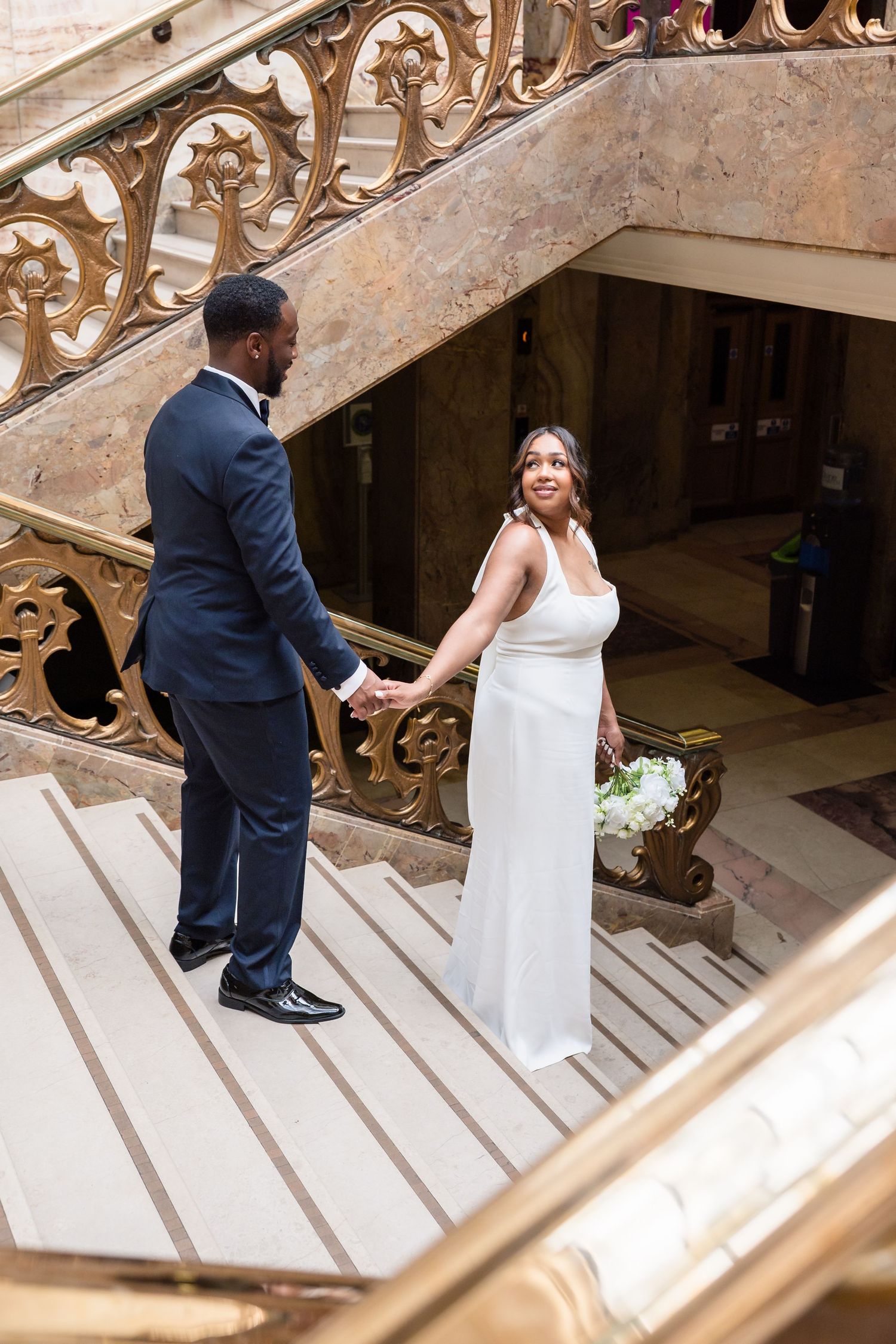 A couple in wedding attire hold hands while standing on elegant ornate stone stairs during their special day.
