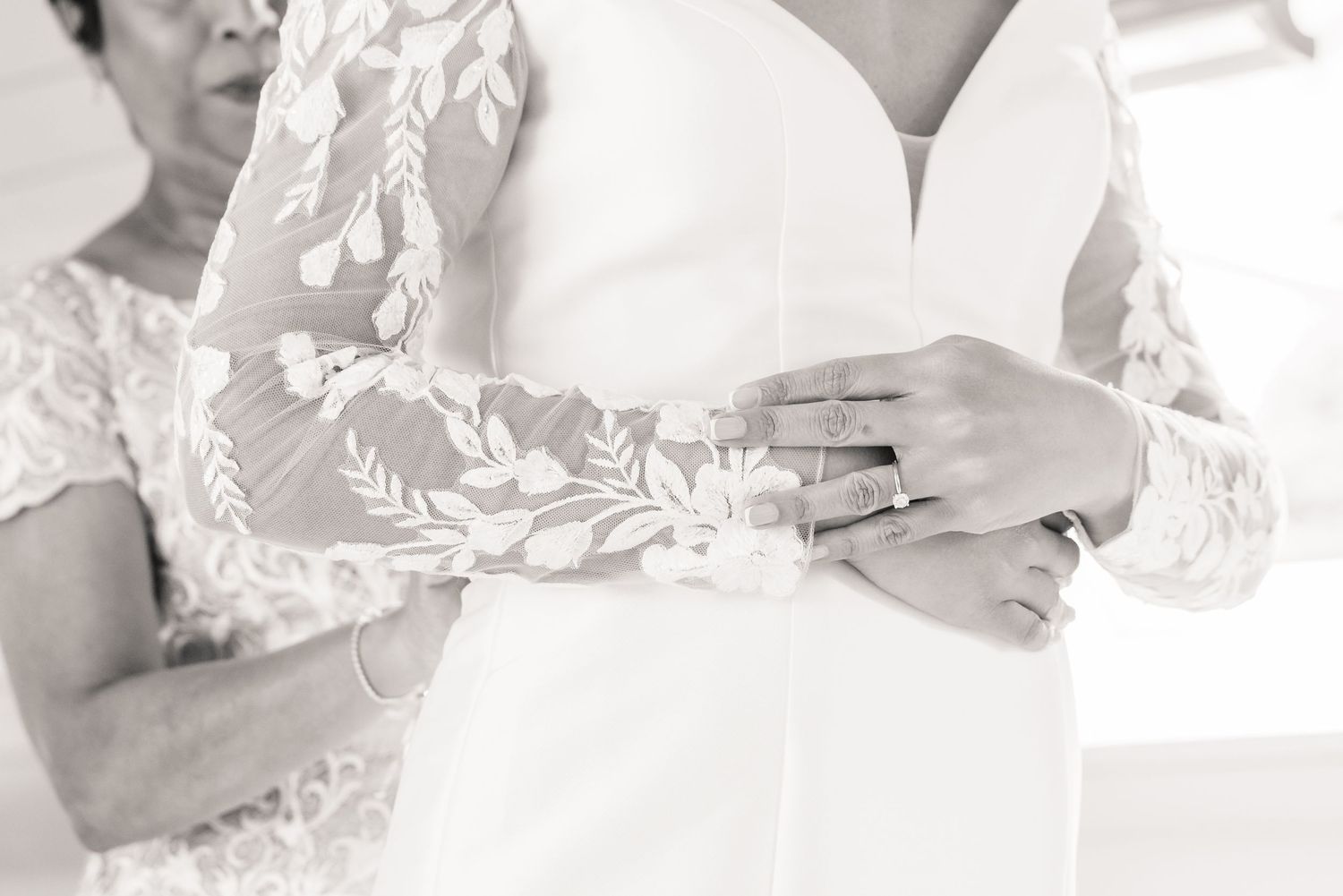 A close up black and white photo of elegant lace wedding dress details and a ring on someone's hand.