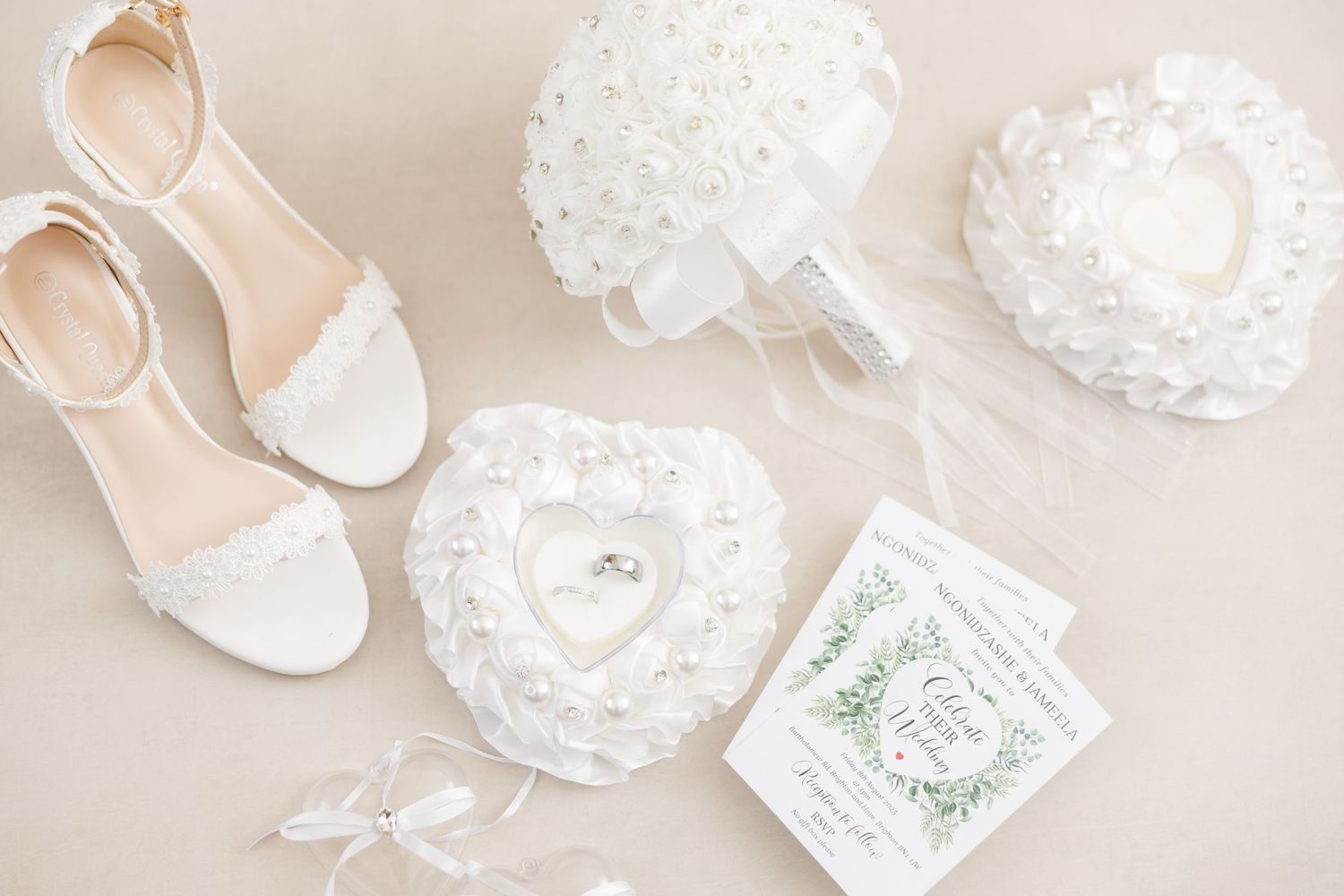 Flat lay of delicate white wedding accessories including lace flowers, shoes, and heart-shaped decorations on beige background.