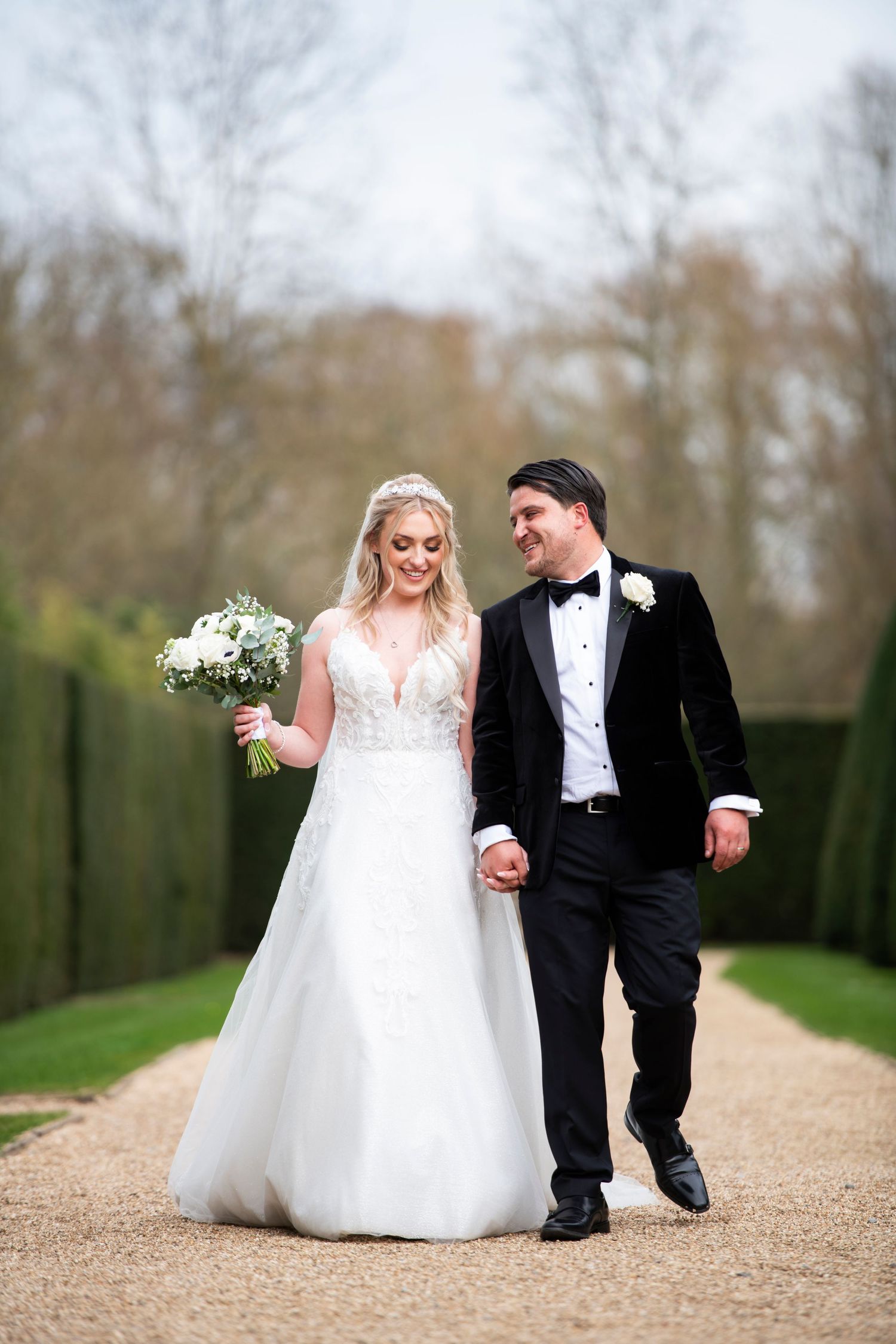 A couple walks hand in hand on a garden path after their wedding ceremony, with her carrying a white floral bouquet.