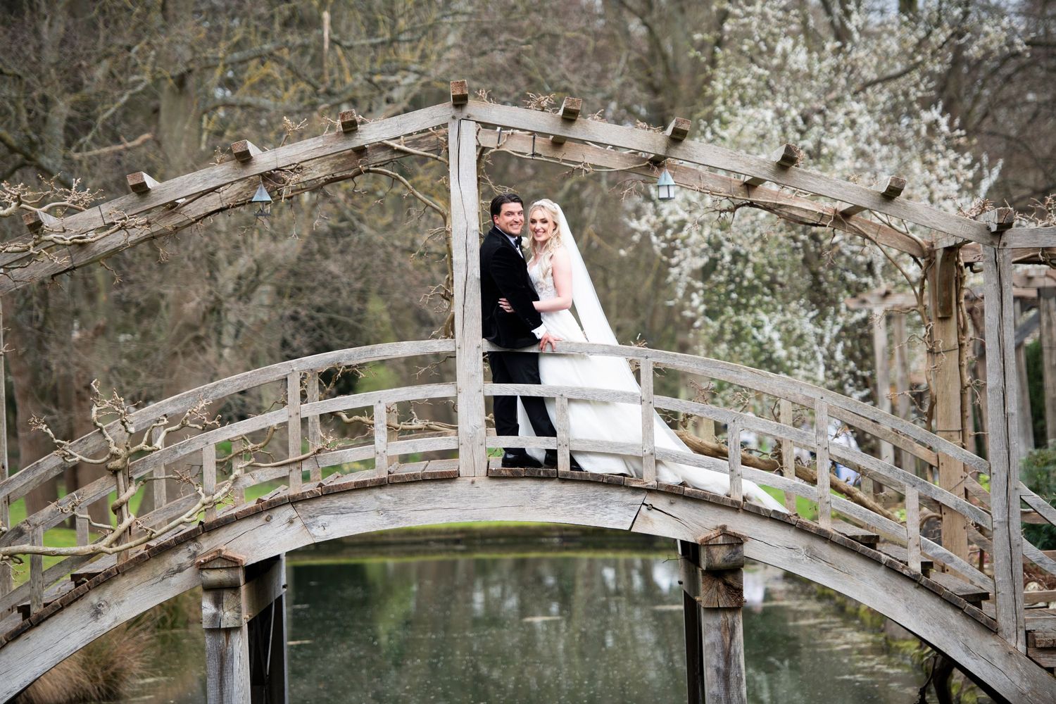 A couple in wedding attire stands together on a rustic wooden footbridge over tranquil water.