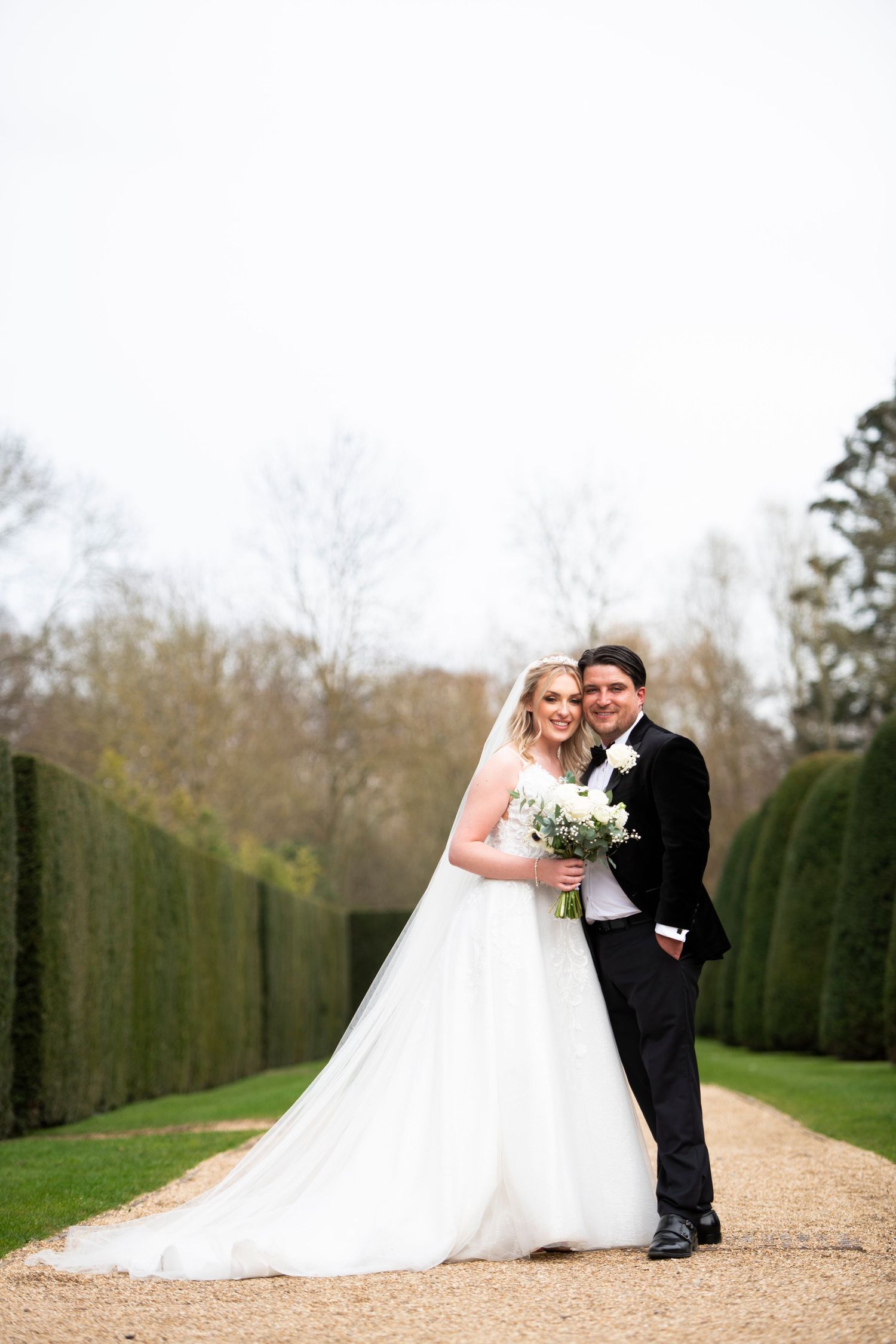 A romantic wedding couple poses on a gravel path lined with manicured hedges in an elegant garden setting.