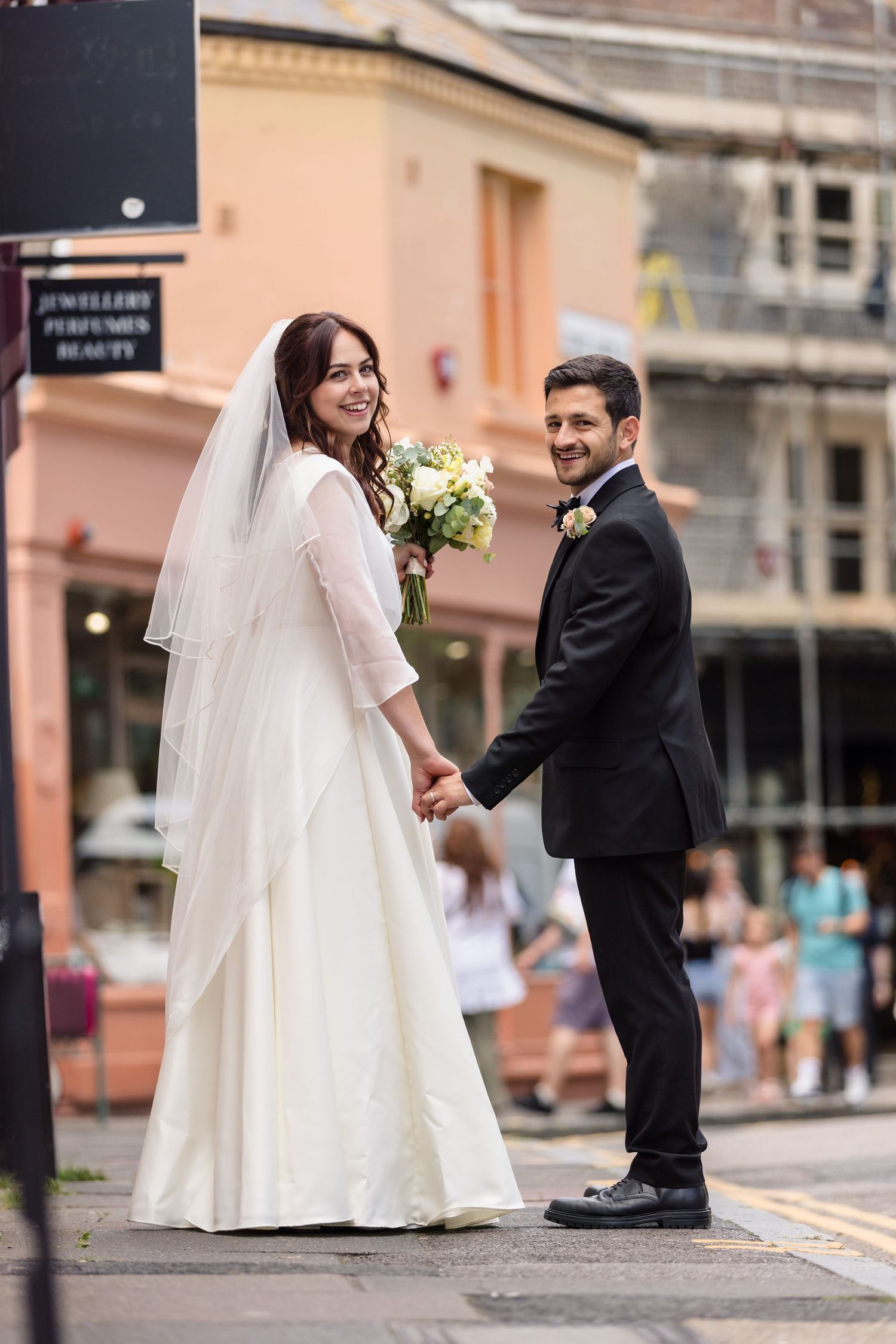 A bride in a white lace wedding dress and groom in black tux hold hands on a city street with pink buildings behind them.