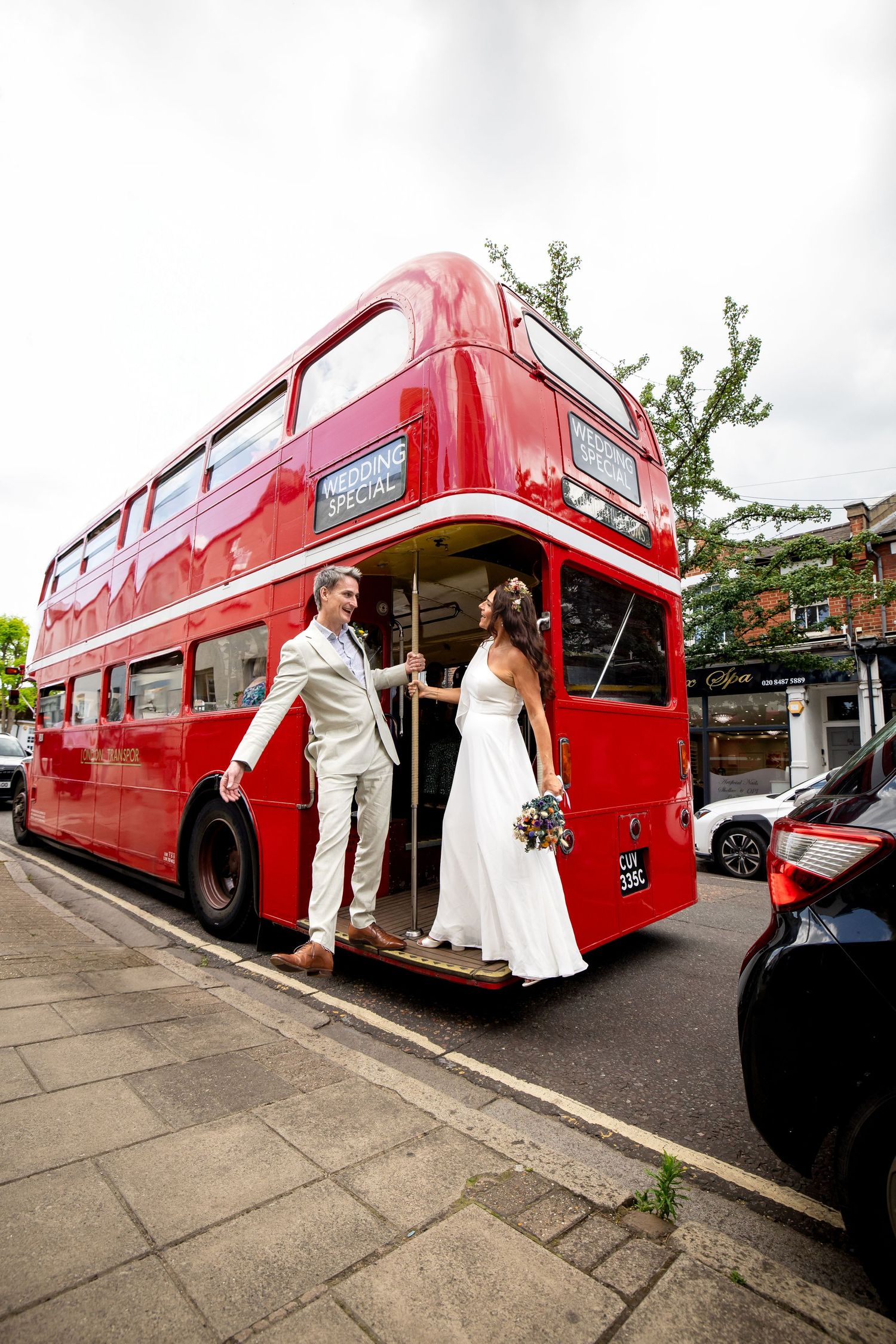 A couple in wedding attire board a classic red London double-decker bus on a city street.