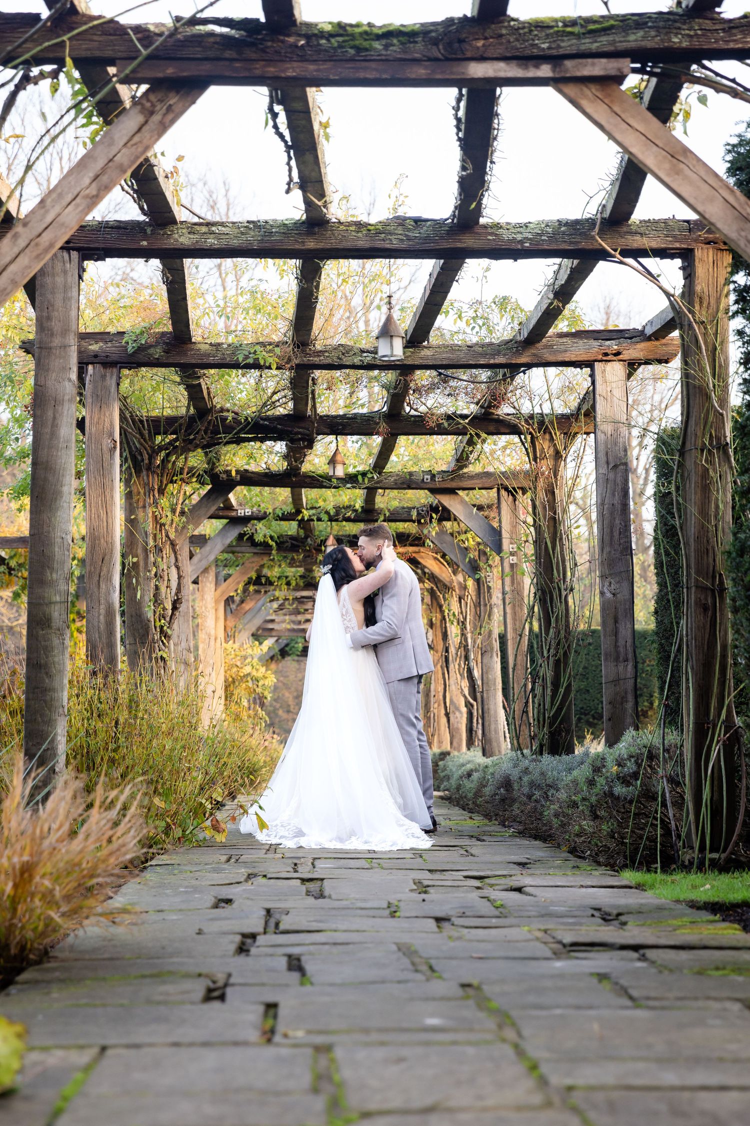 Wedding couple embracing under a rustic wooden pergola in a garden setting.