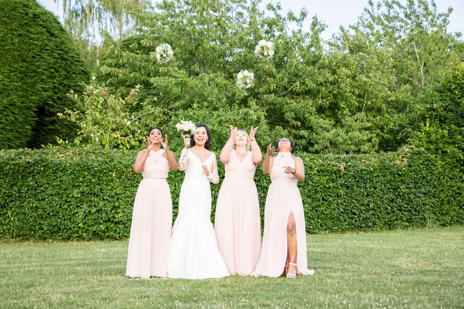 A line of bridesmaids in blush pink dresses pose together on a lush green lawn with hedges in the background.