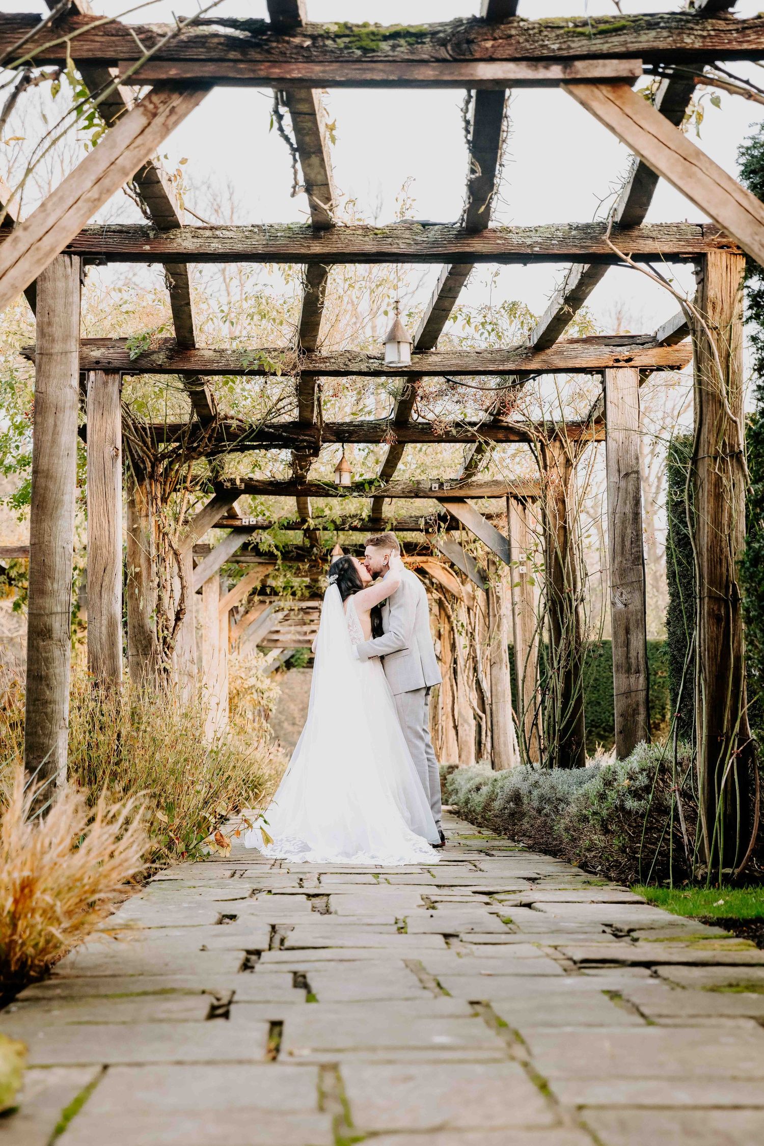A romantic wedding moment under a rustic wooden pergola covered in vines along a stone garden path.