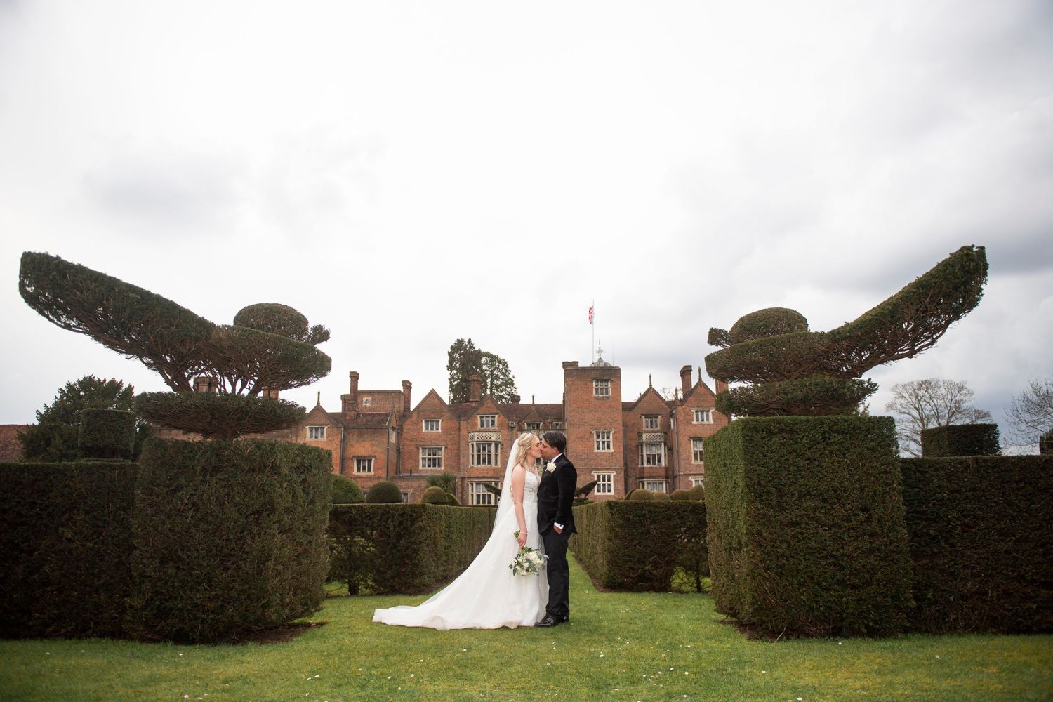 Wedding couple stands between ornate topiary birds at elegant brick manor house with manicured hedges.