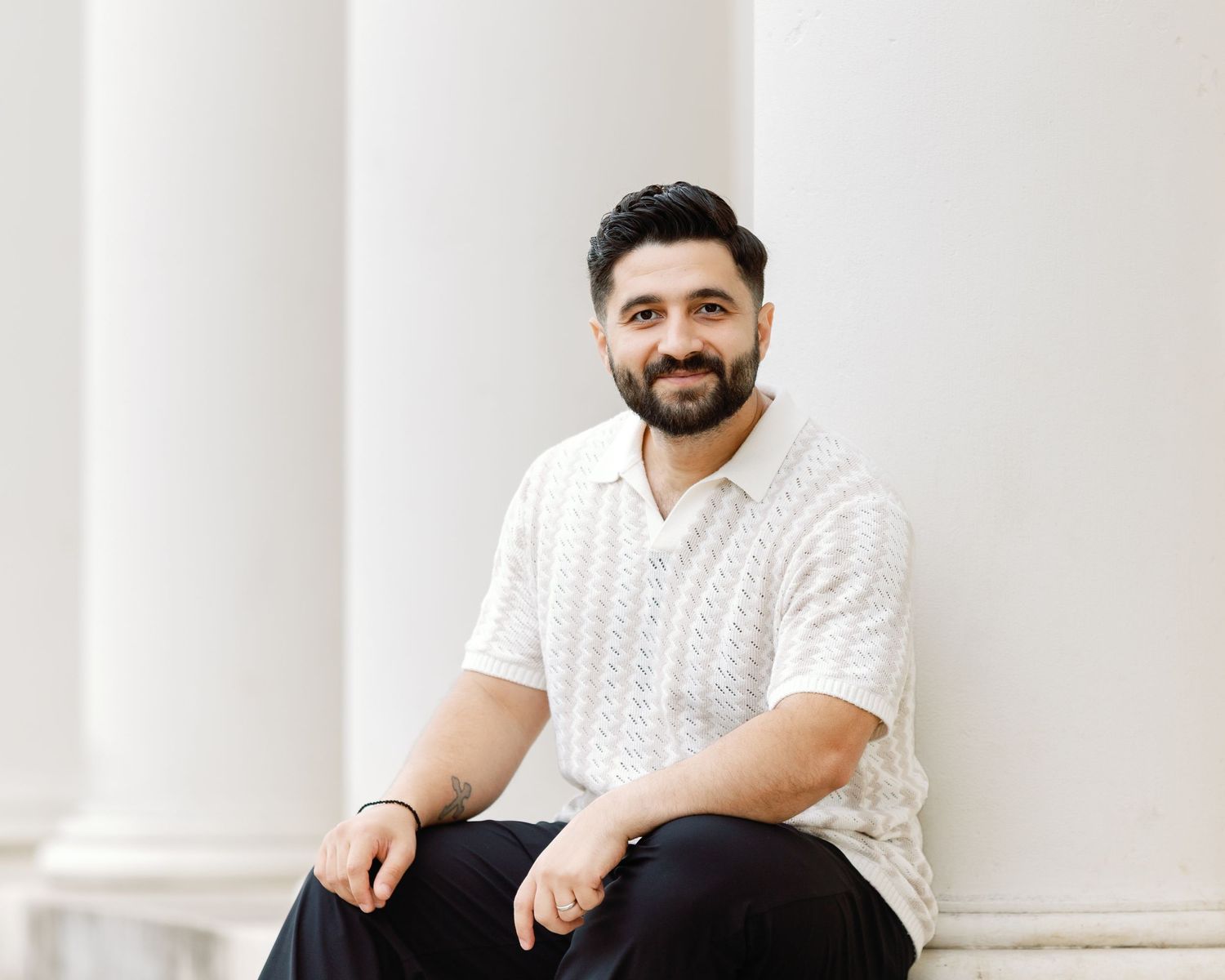 A person in a white cable knit sweater smiles while sitting casually against a white column background.