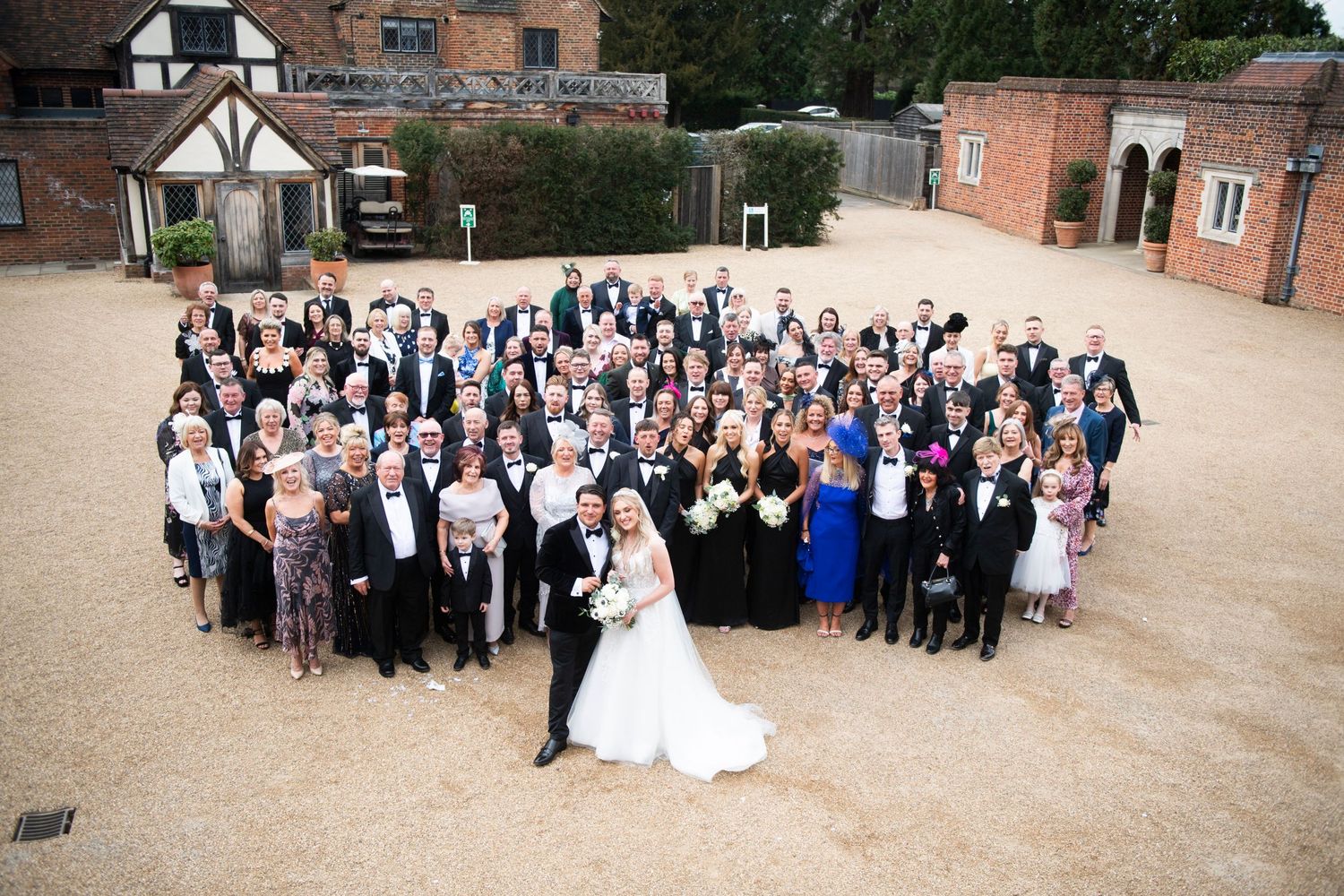 Large outdoor wedding party group photo taken from above on a sandy courtyard with brick buildings in the background.