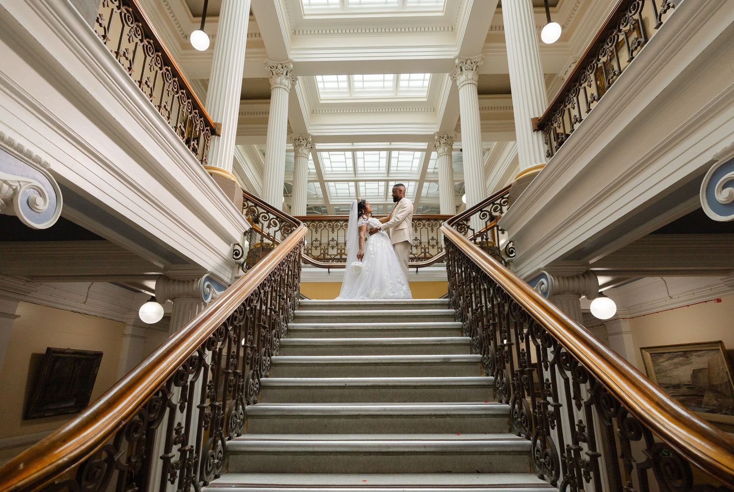 Elegant grand staircase with ornate iron railings and wooden banisters leads up to a skylit landing in historic building.