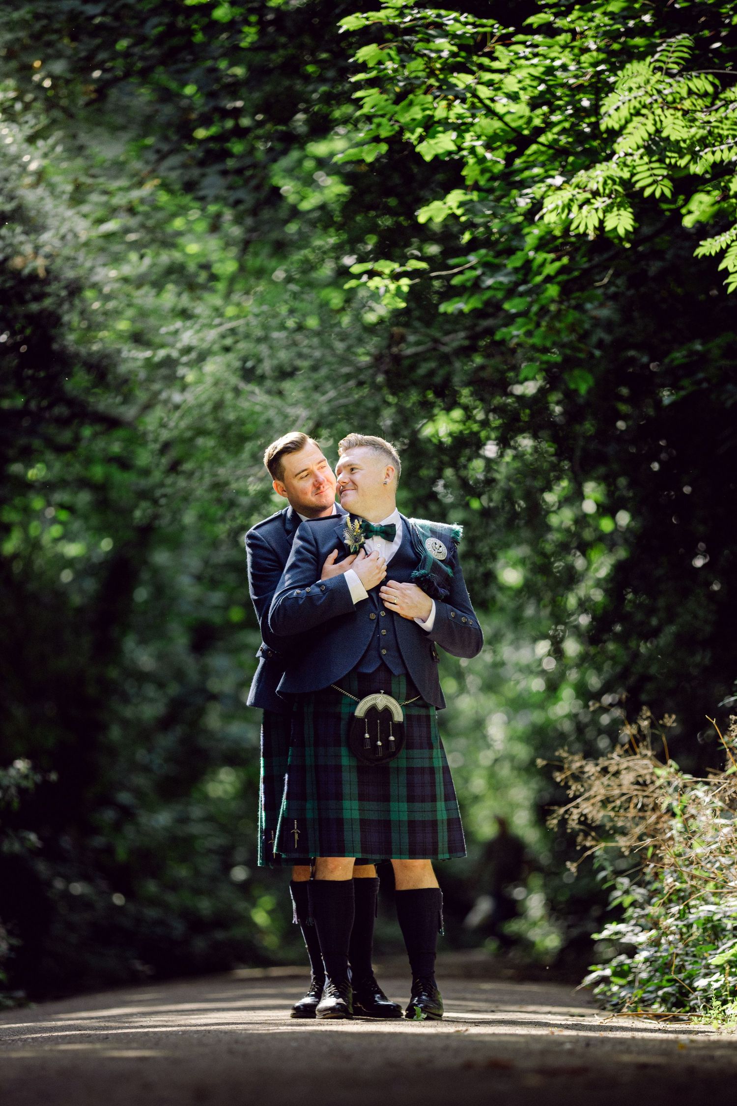 Two people in traditional Scottish kilts and formal jackets share a romantic moment on a tree-lined path.