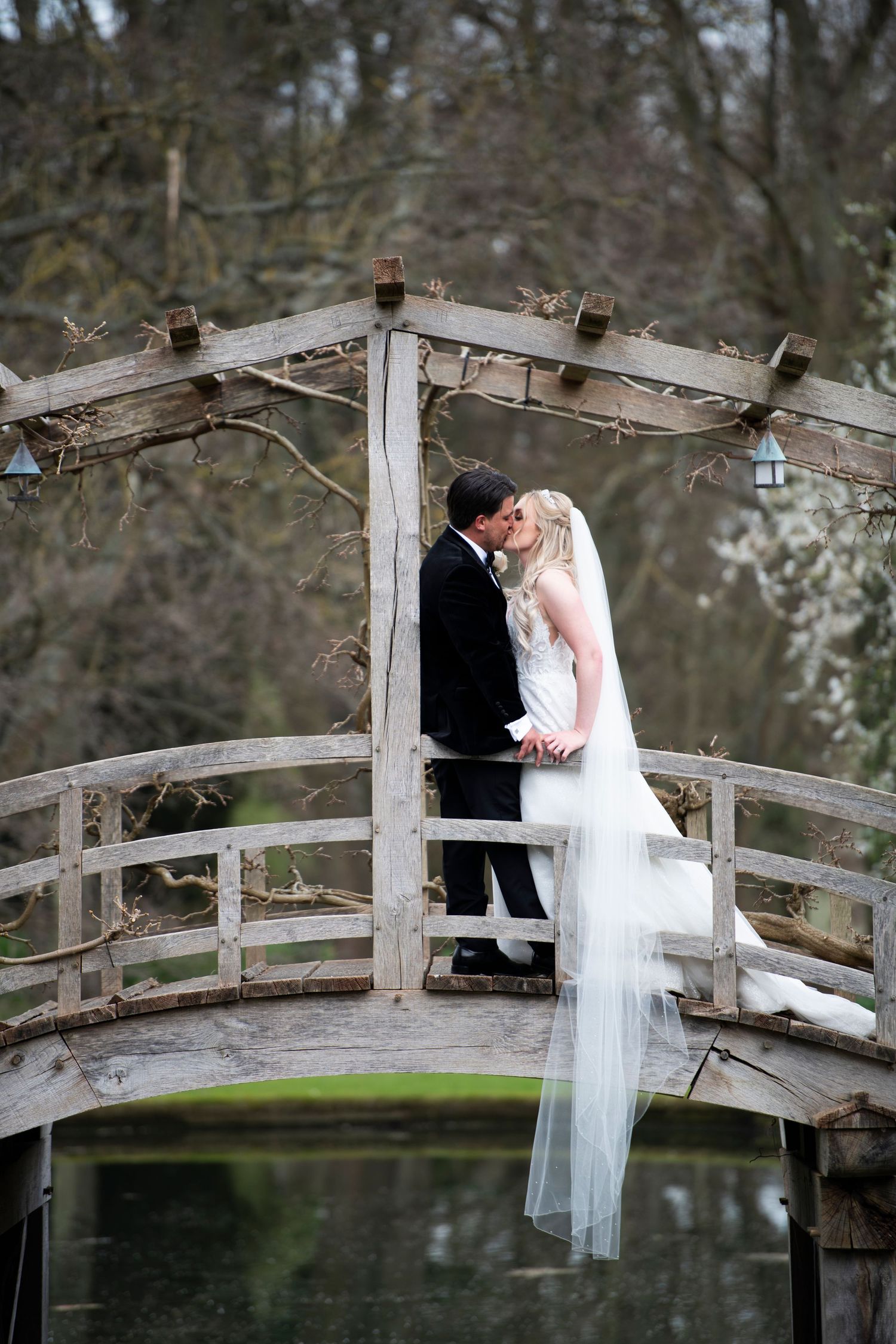 A wedding couple shares an intimate moment on a rustic wooden footbridge over a pond in a romantic garden setting.