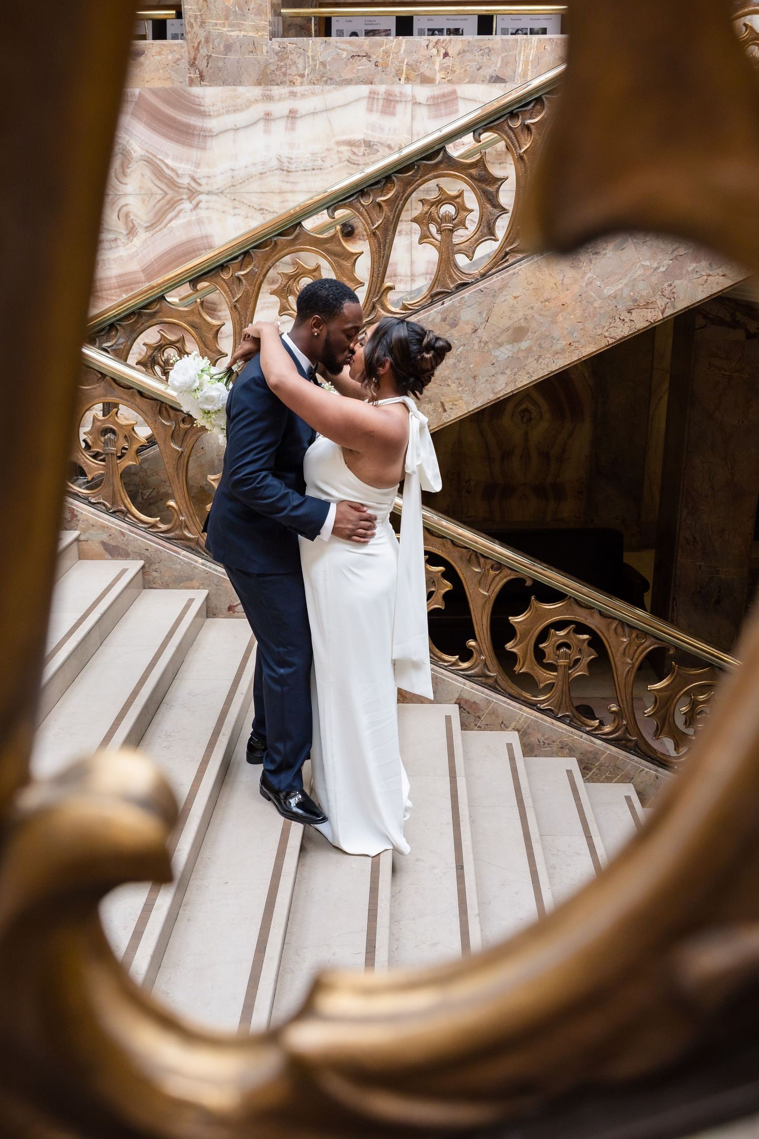 Couple in navy suit and white gown embrace on an ornate vintage staircase with decorative metal railings.