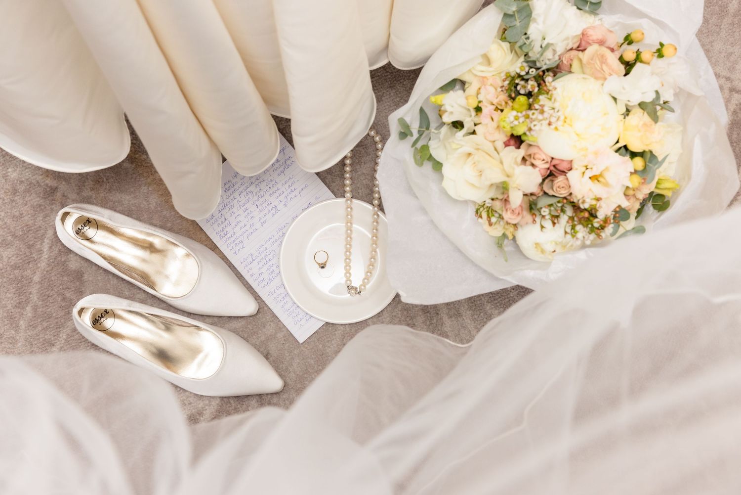 White bridal shoes and delicate cream-colored bouquet with roses and greenery arranged on light fabric.