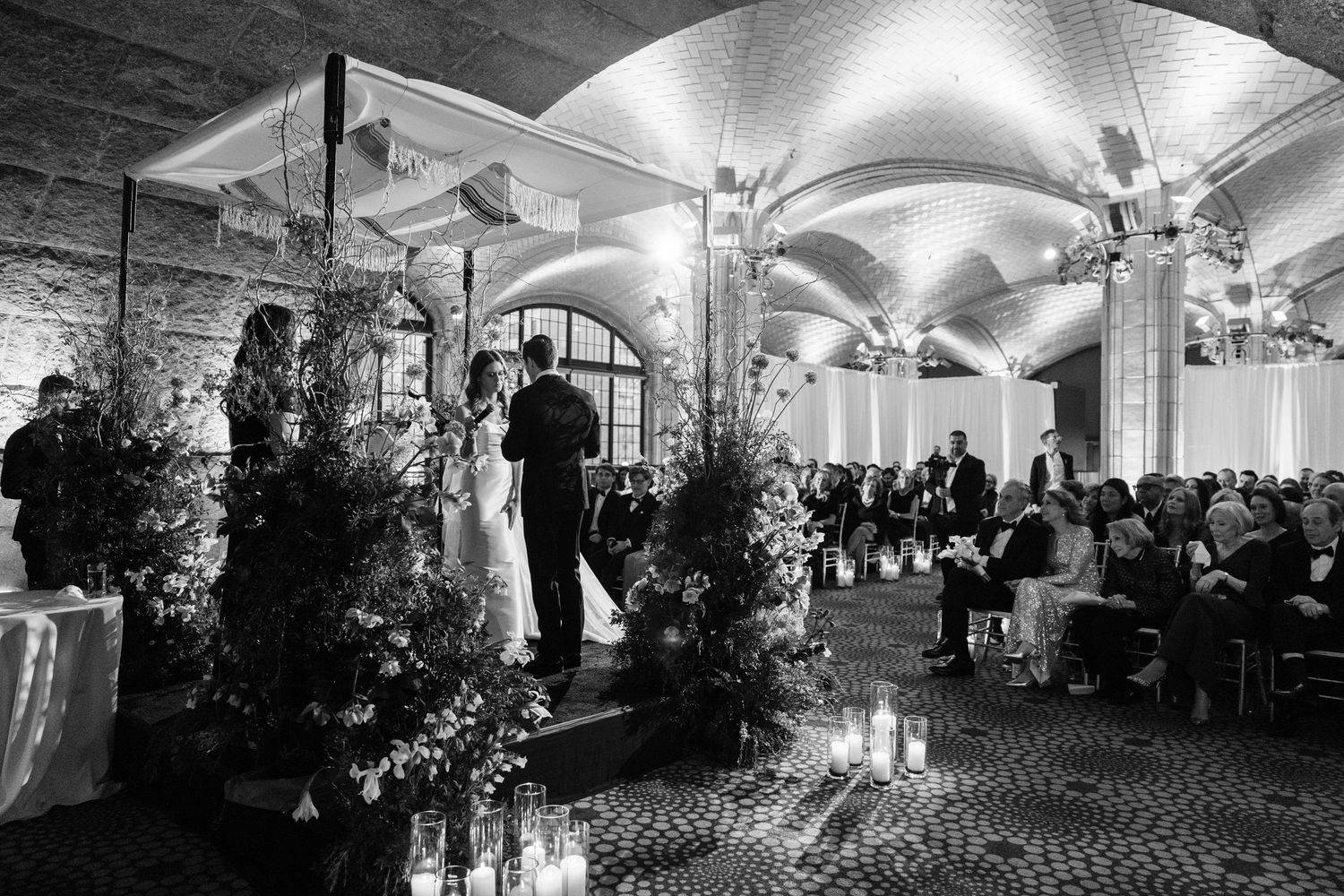 Black and white wedding ceremony in ornate vaulted hall with candlelit aisle and seated guests.