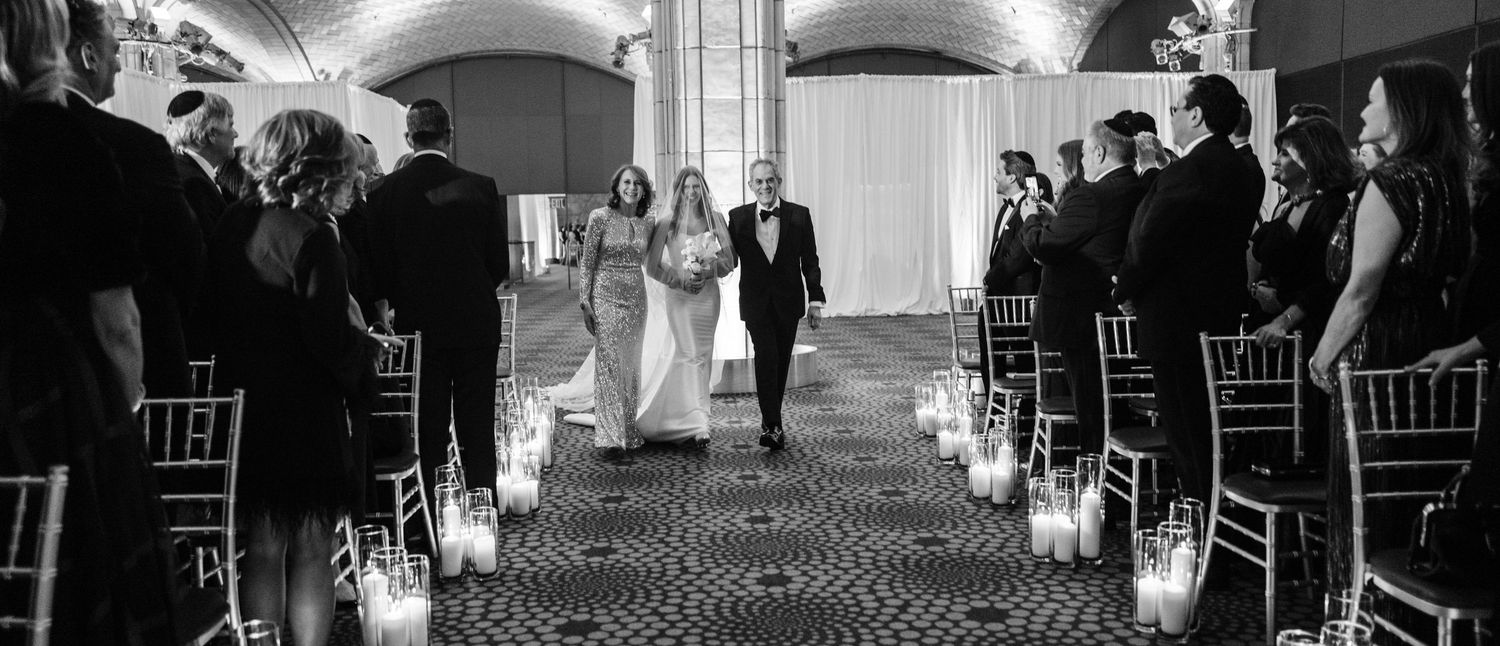 Bride walks down candlelit wedding aisle escorted by parents in elegant venue, black and white photo.