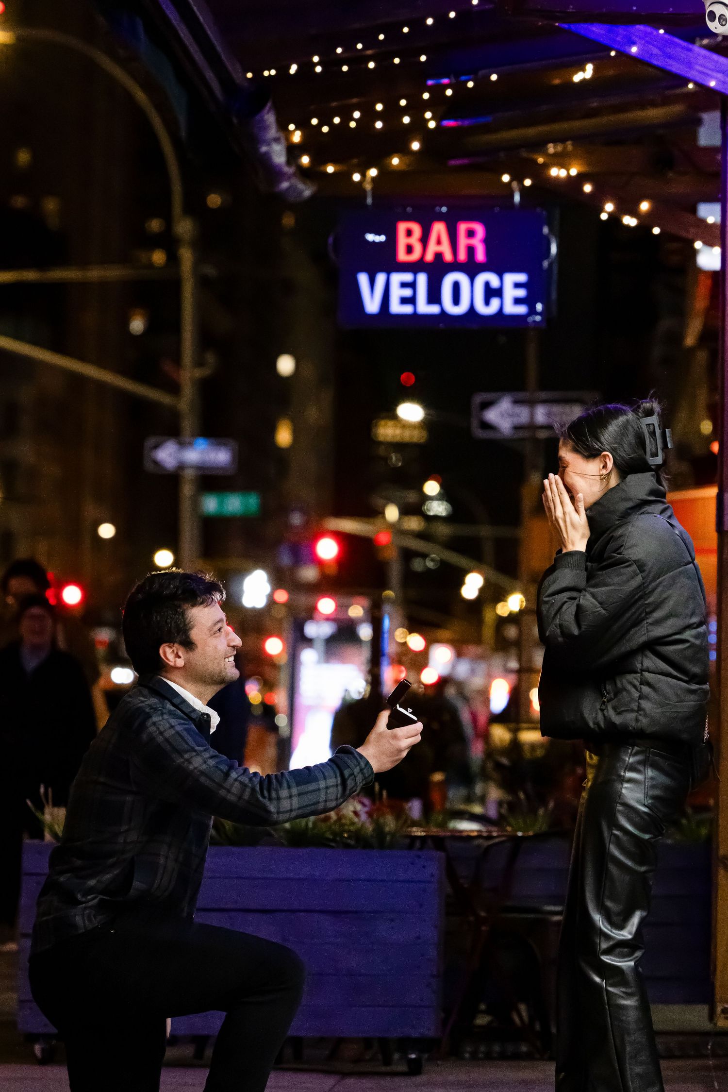 Man proposes on one knee to surprised woman outside Bar Veloce in New York City at night.