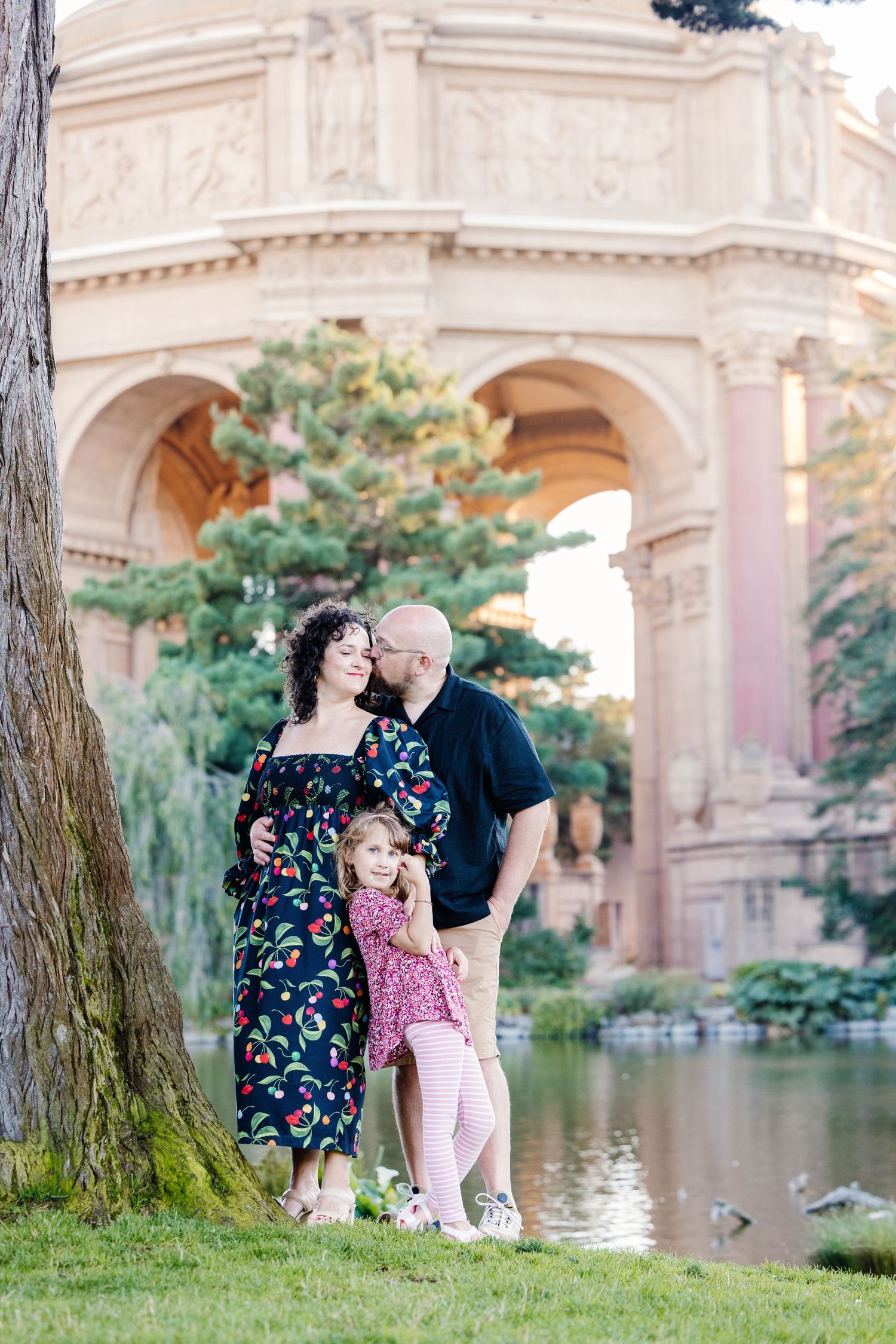 Family of three stands near a pond at the Palace of Fine Arts at golden hour in San Francisco.