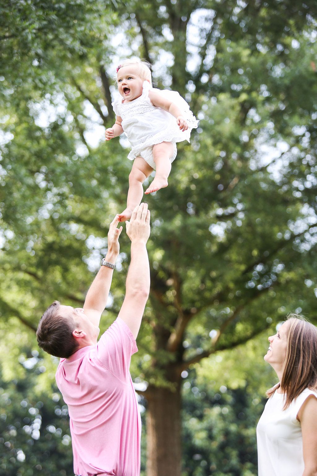 Father tossing baby girl in the air outdoors while mother watches, family fun in a green park setting.