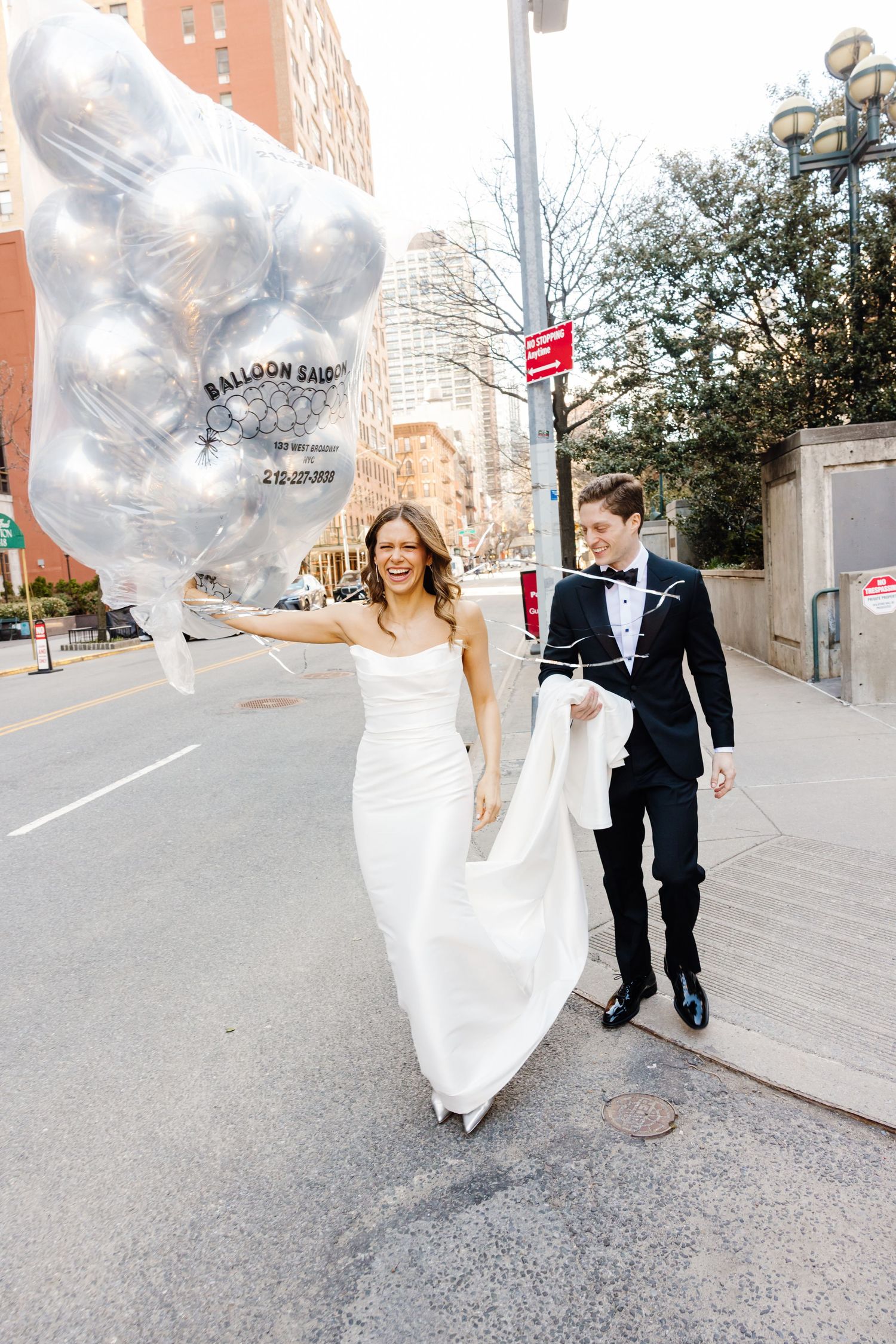 Bride holds silver balloons while walking with groom in tuxedo on New York City street.