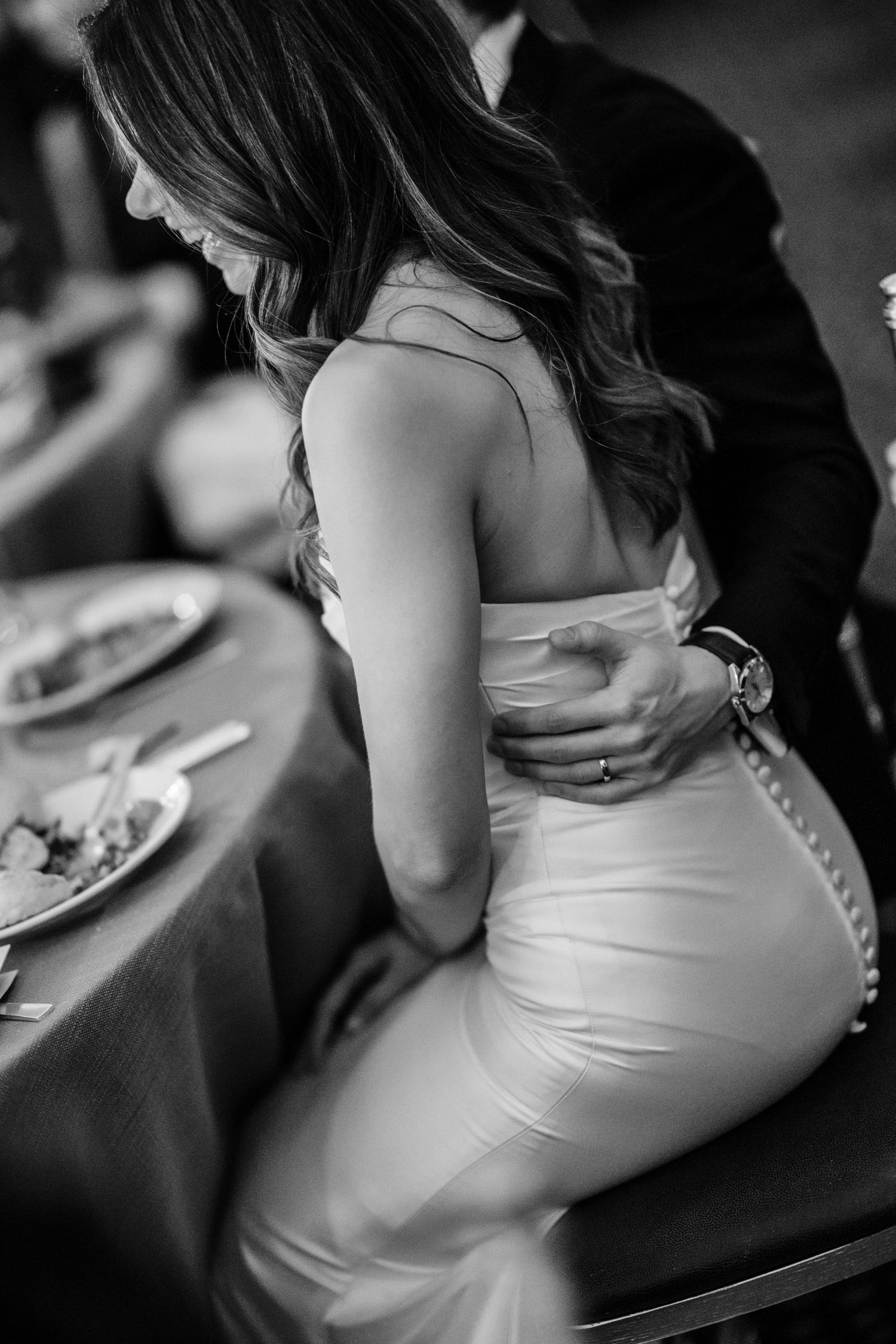 Black and white photo of groom embracing bride in fitted white gown at wedding reception.