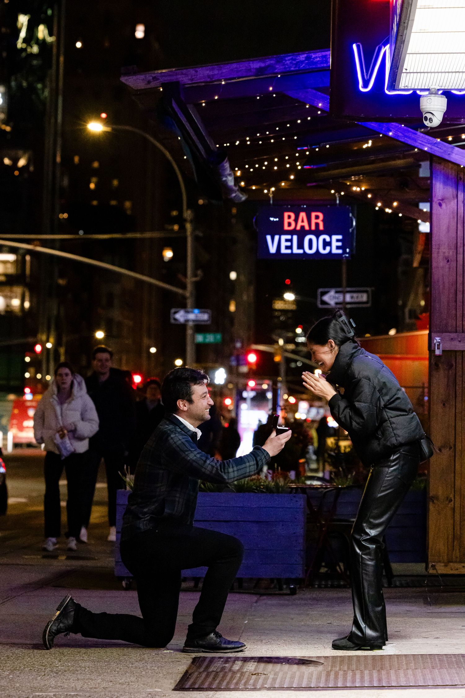 Man proposing on one knee outside Bar Veloce on a New York City street at night.