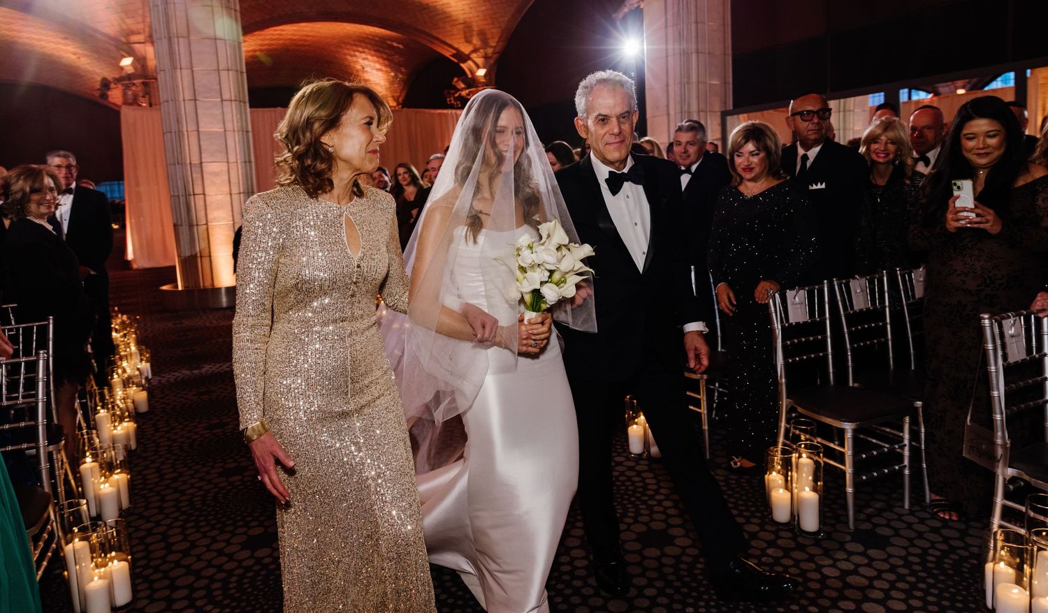Bride in white gown and veil walks down candlelit aisle escorted by parents at elegant wedding.