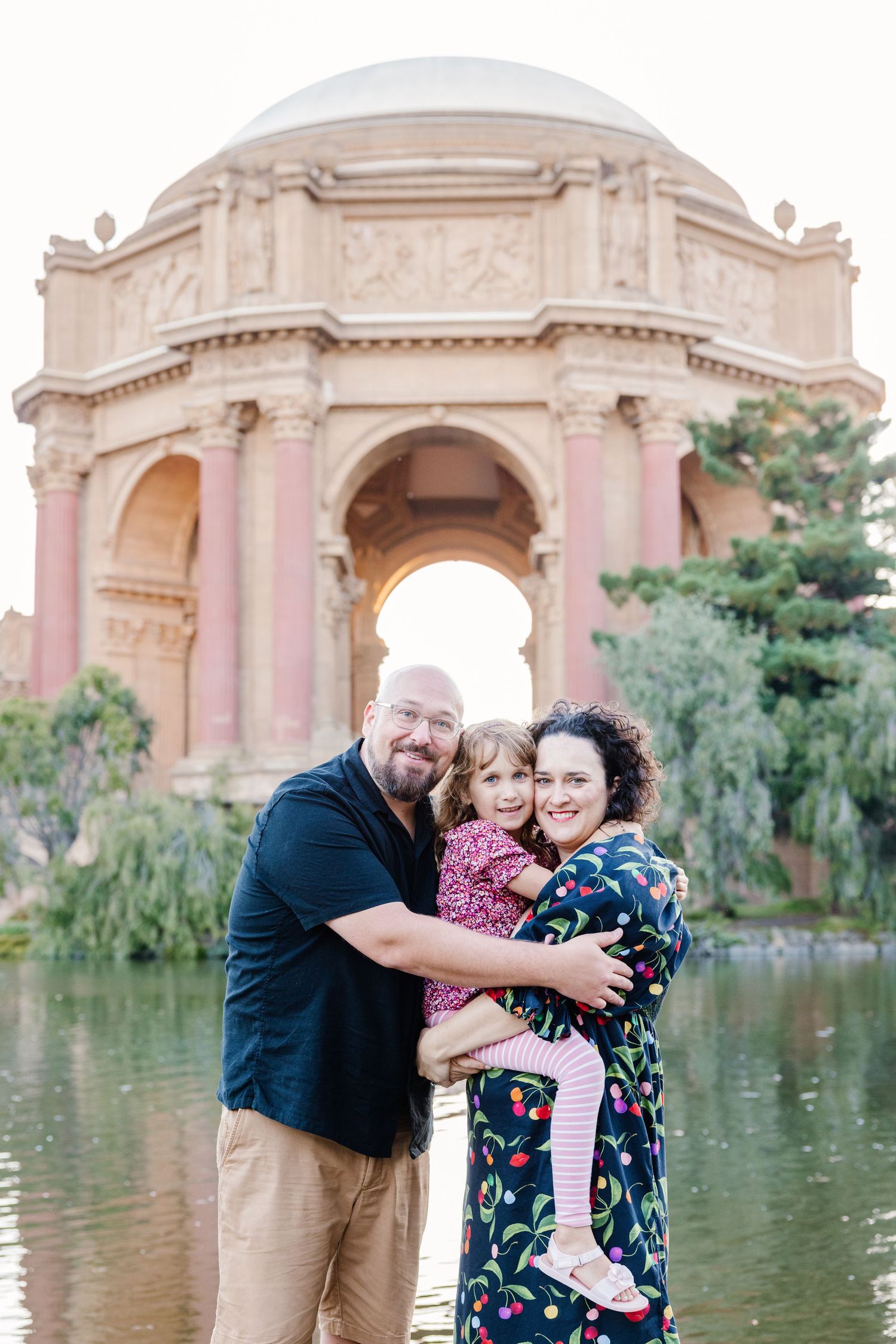 Smiling family of three hugs in front of the Palace of Fine Arts rotunda at sunset.