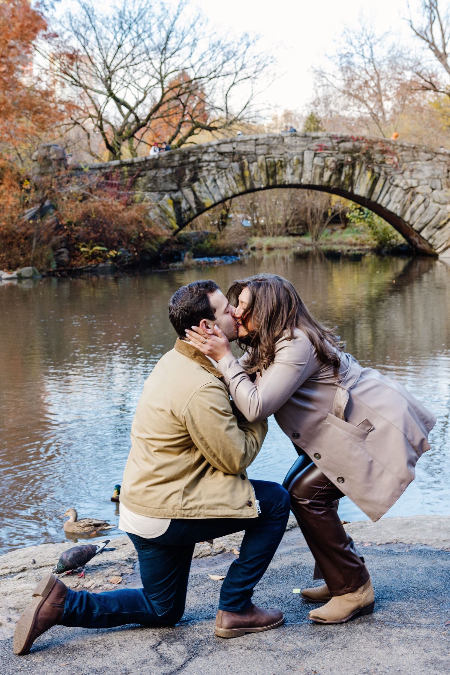 Couple sharing a kiss after proposal near Gapstow Bridge in Central Park during autumn.