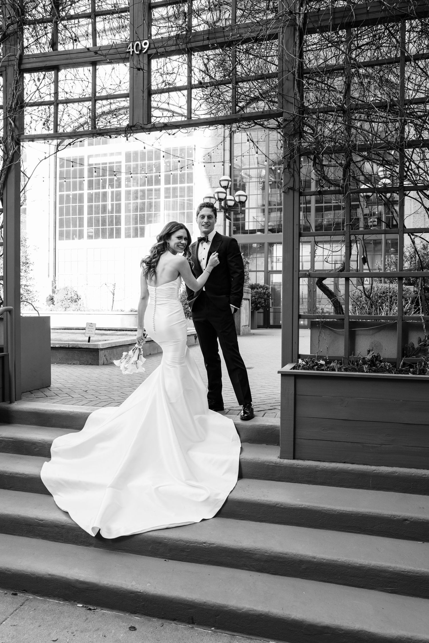Black and white photo of bride and groom posing on steps outside a building with a long train.