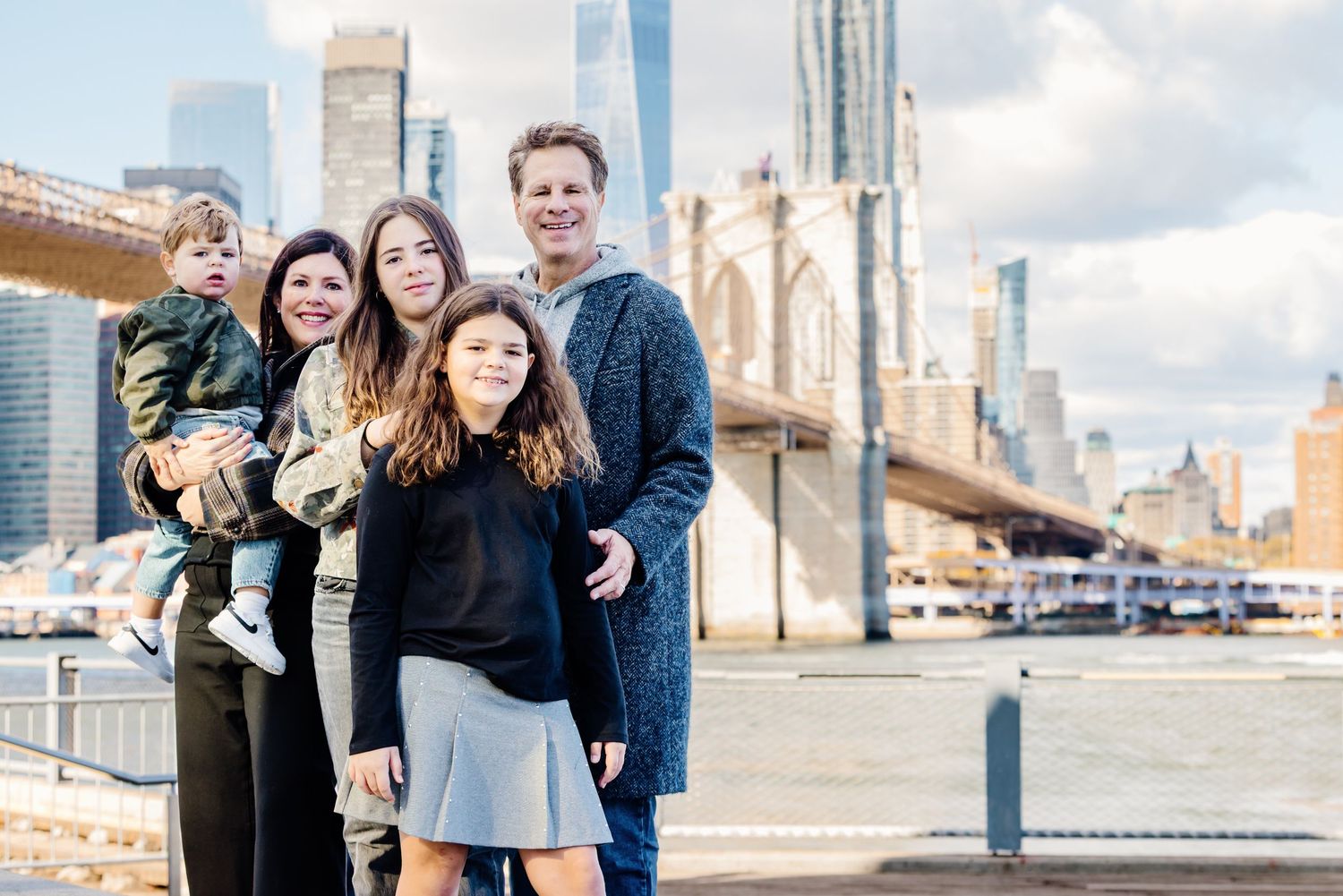 Happy family of five poses by the East River with Brooklyn Bridge and NYC skyline behind them.