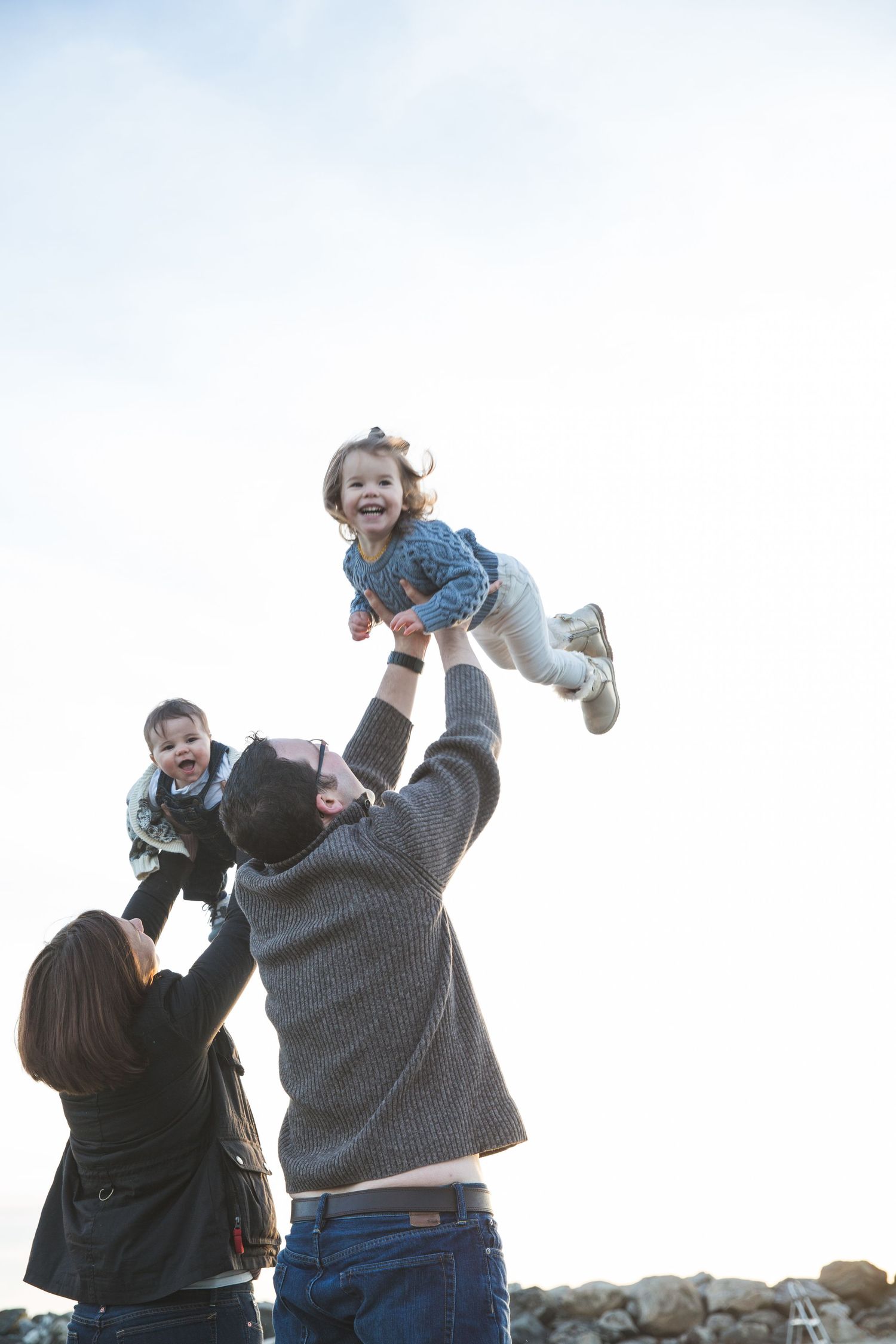 A joyful family outdoors, father lifting laughing toddler high in the air while mother holds baby against a bright sky.