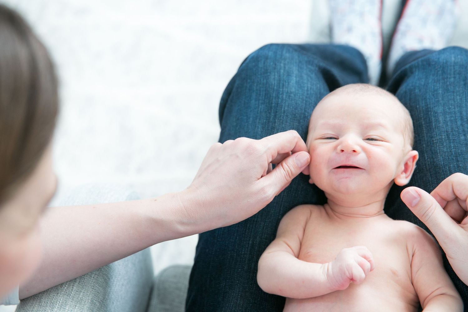Smiling newborn baby being gently touched on the cheek by a parent while resting on an adult's lap.