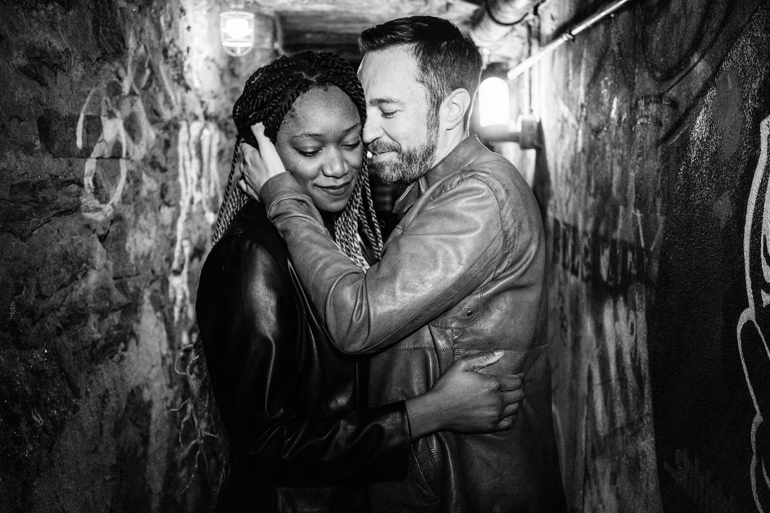 Black and white photo of an interracial couple embracing and laughing together in a graffiti-covered alley.