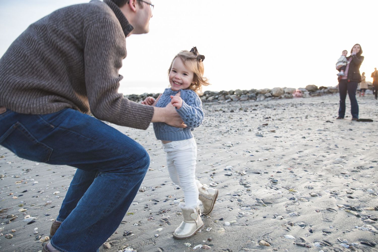 Father crouching on a rocky beach, smiling at his laughing toddler girl in white pants and bow headband.