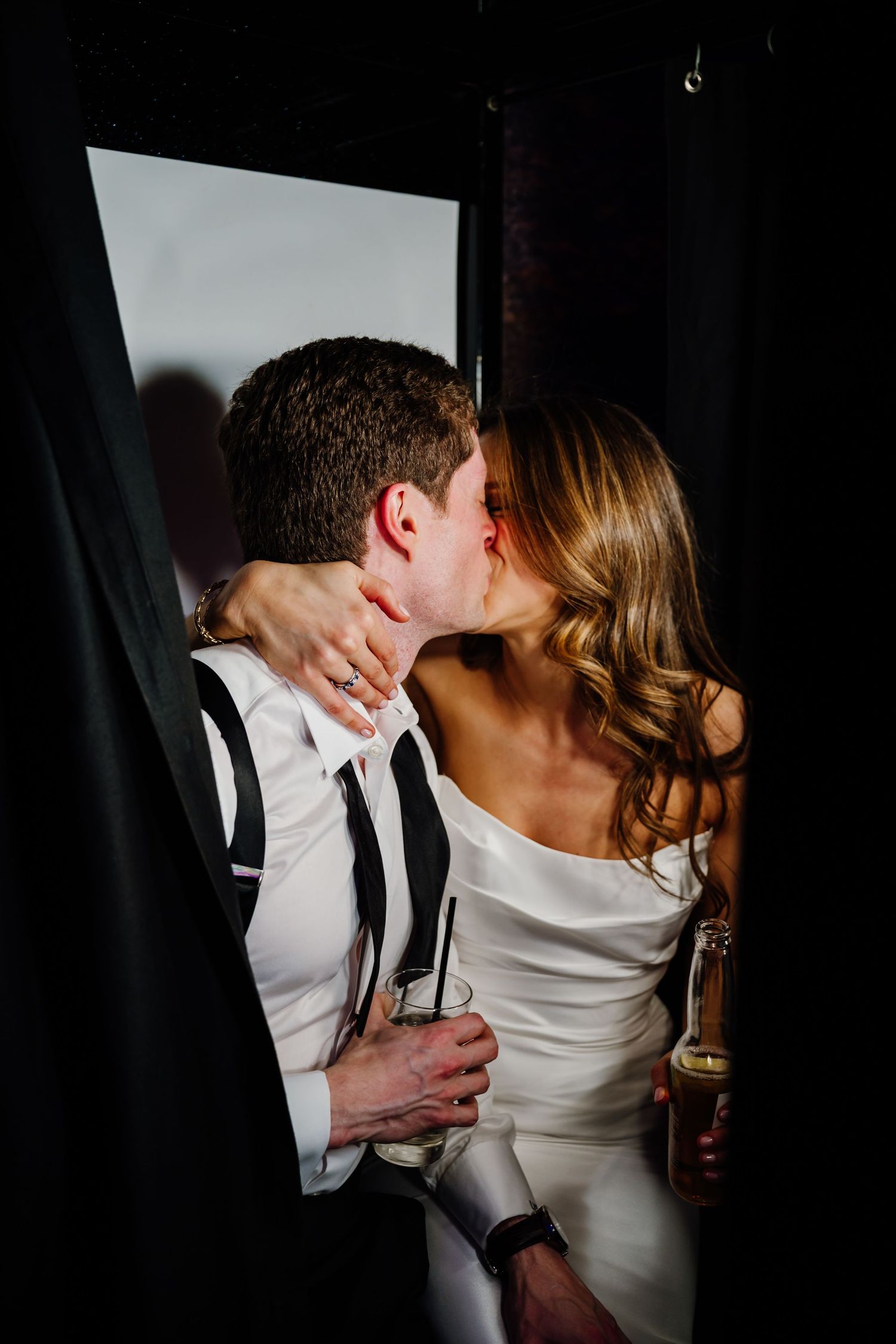 Newlyweds kissing passionately in photo booth while holding champagne at wedding.