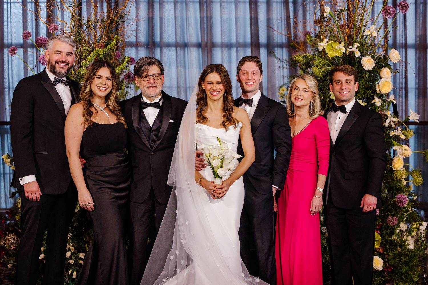 Wedding party of seven posing in formal attire before floral backdrop at elegant indoor ceremony.