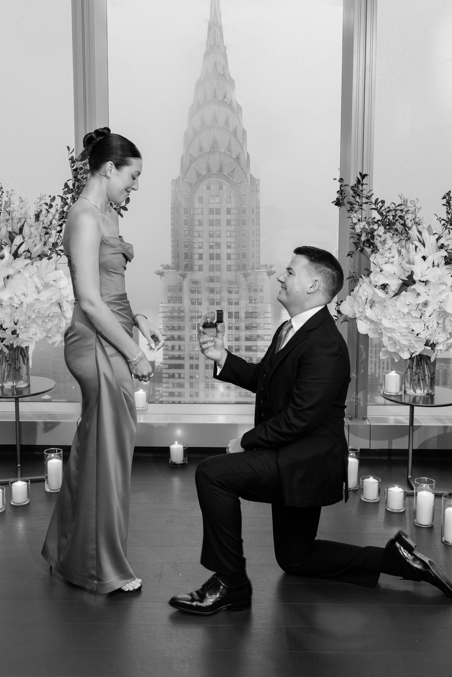 Black and white photo of man proposing on one knee with Chrysler Building visible through window behind them.