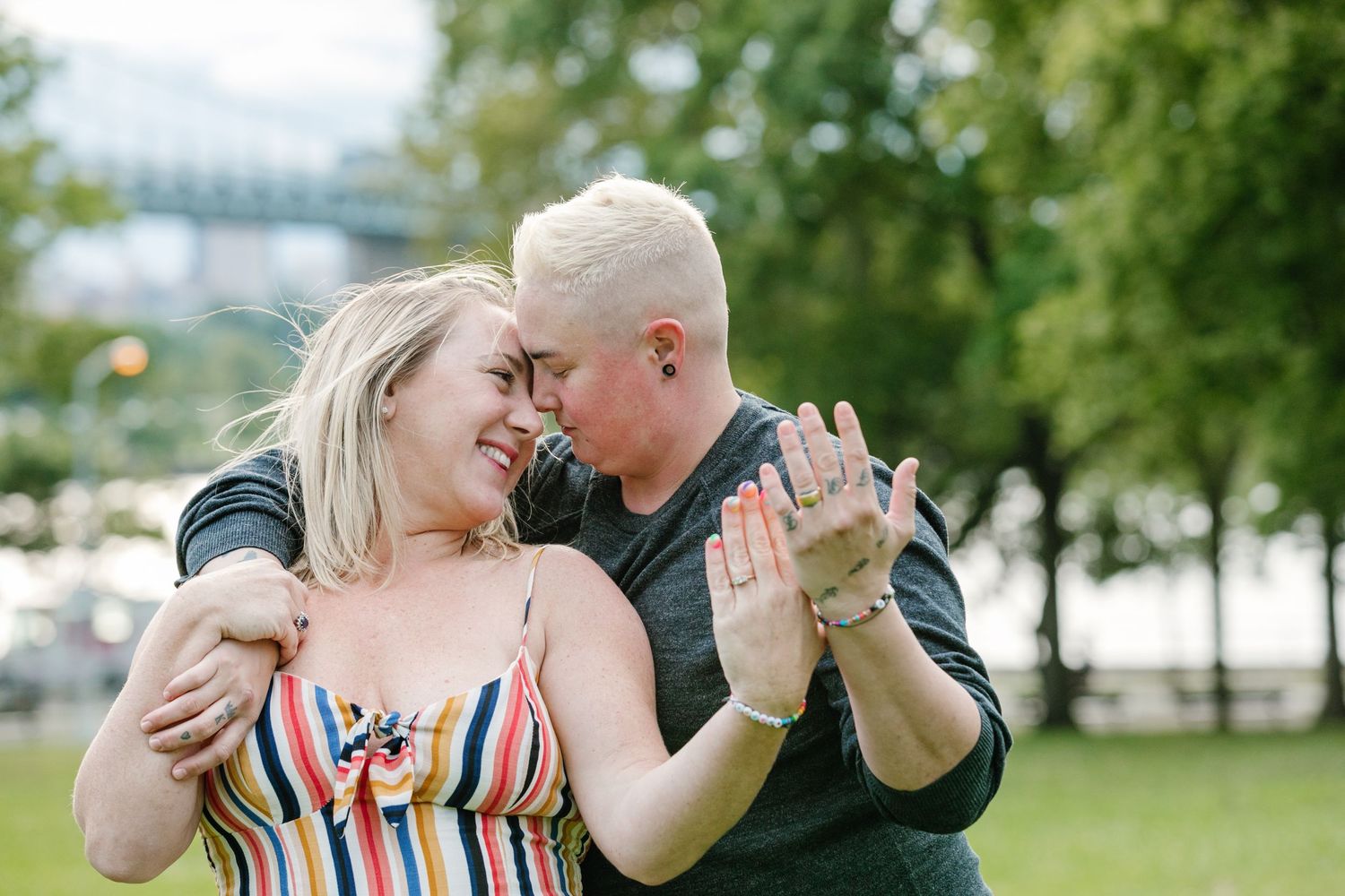 Two women embrace and touch foreheads while dancing outdoors in a park with a bridge visible in the background.