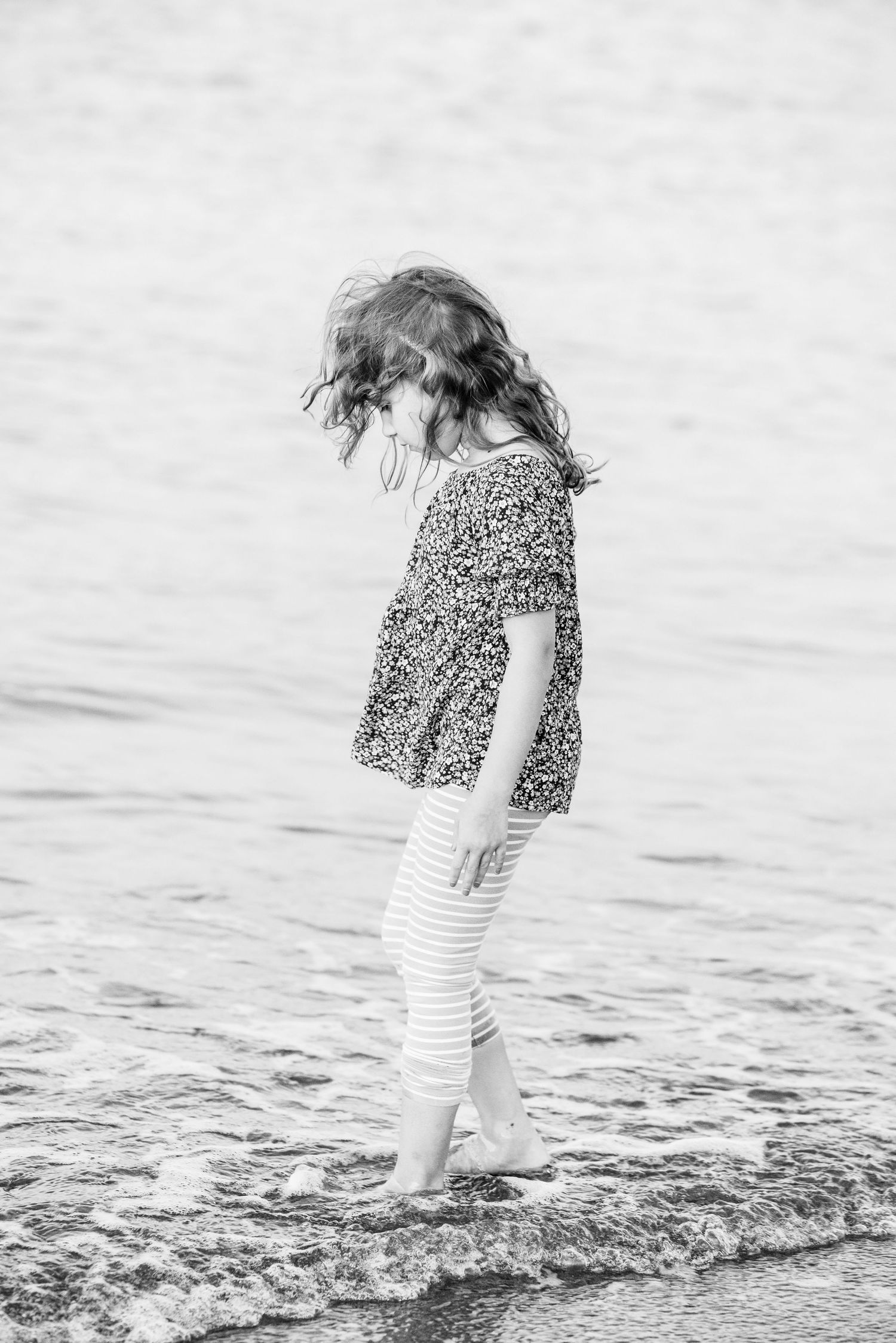 Black and white photo of a young girl in floral top and striped leggings wading barefoot in ocean waves at the beach.