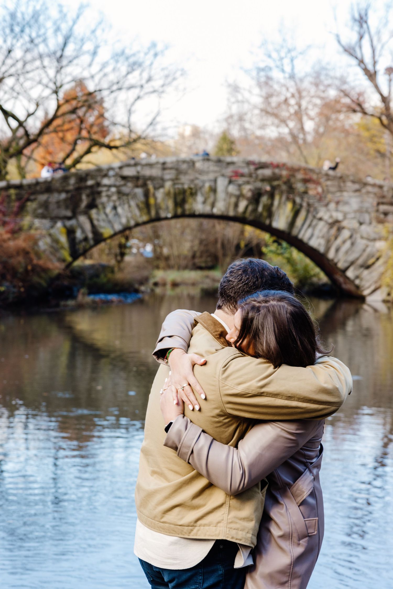 Couple embracing tenderly in front of a stone arch bridge over a calm river during autumn.