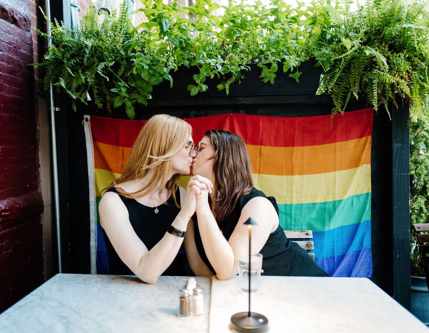 Two women kissing at an outdoor table in front of a rainbow Pride flag.