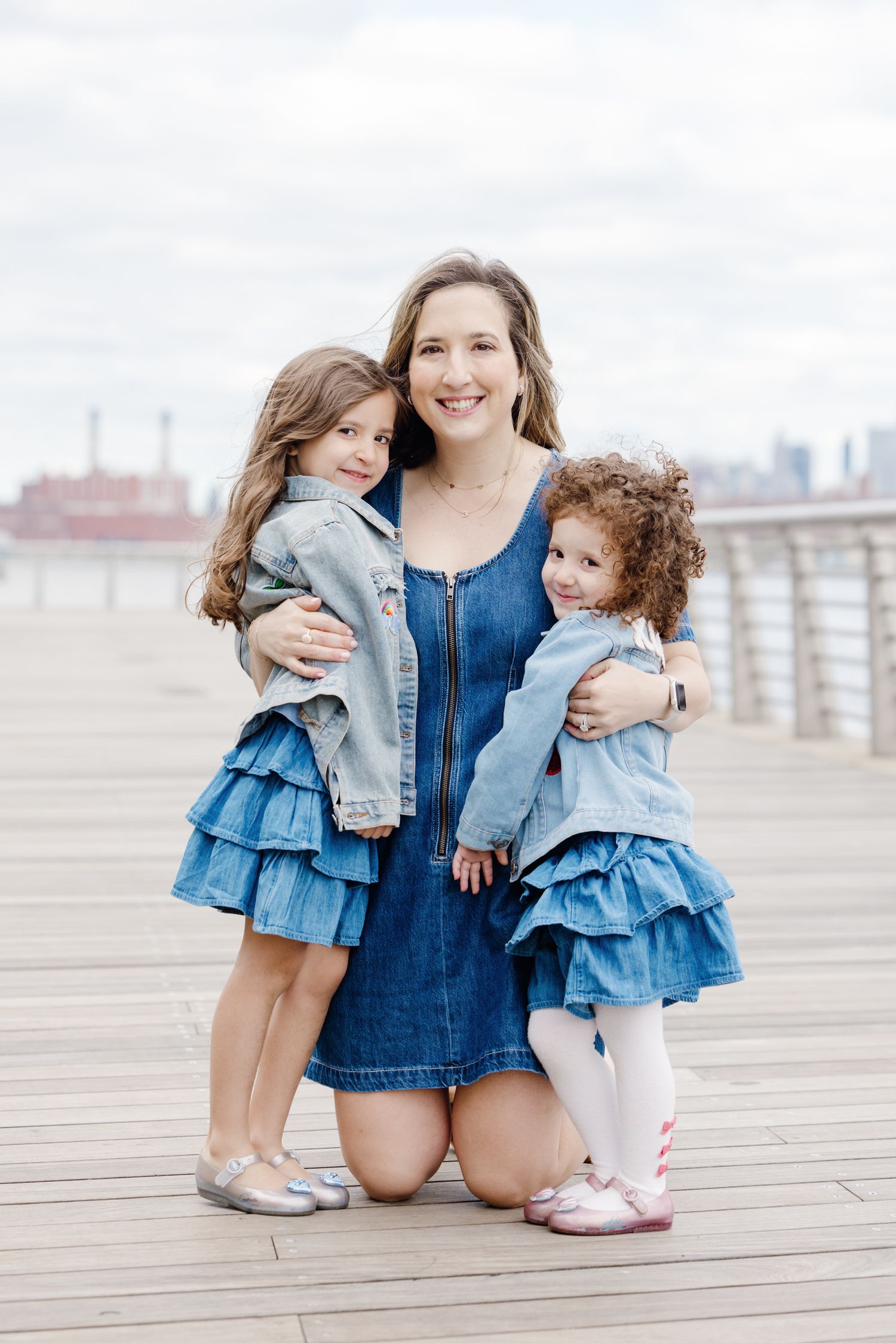 Mother kneeling on boardwalk hugging two young daughters in matching denim outfits by the water.