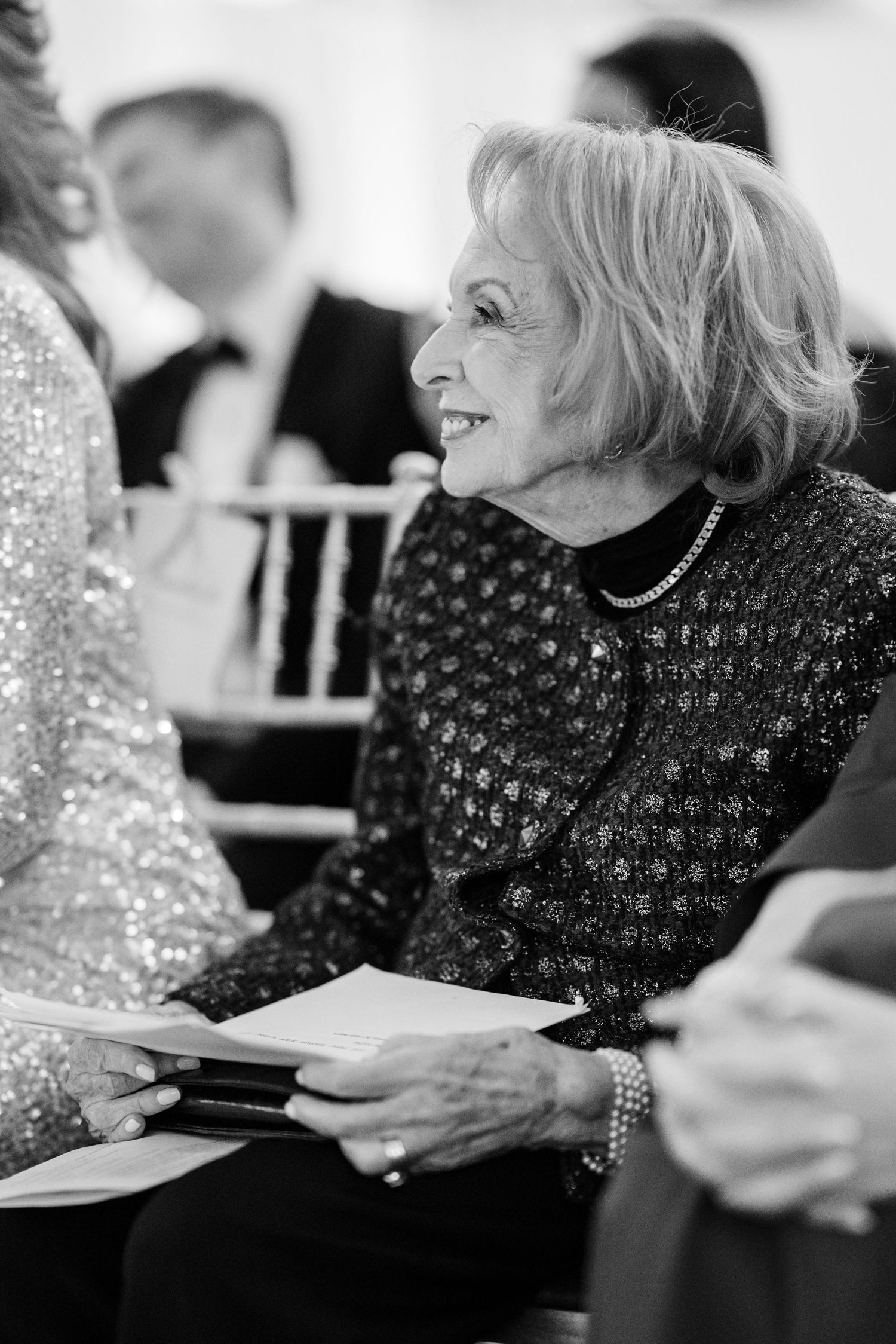 Elegant elderly woman in sequined jacket smiles warmly while holding wedding program at ceremony.