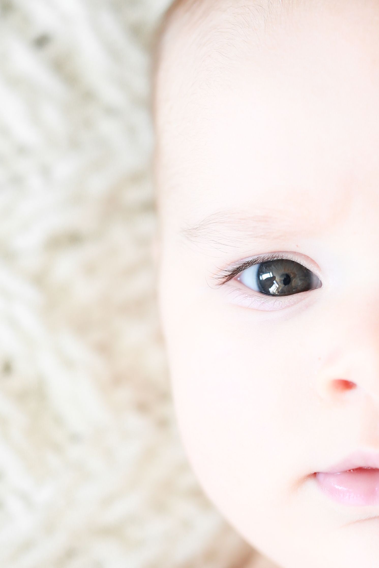 Close-up portrait of a baby's face with bright blue-gray eyes, soft skin, and delicate features on a light background.
