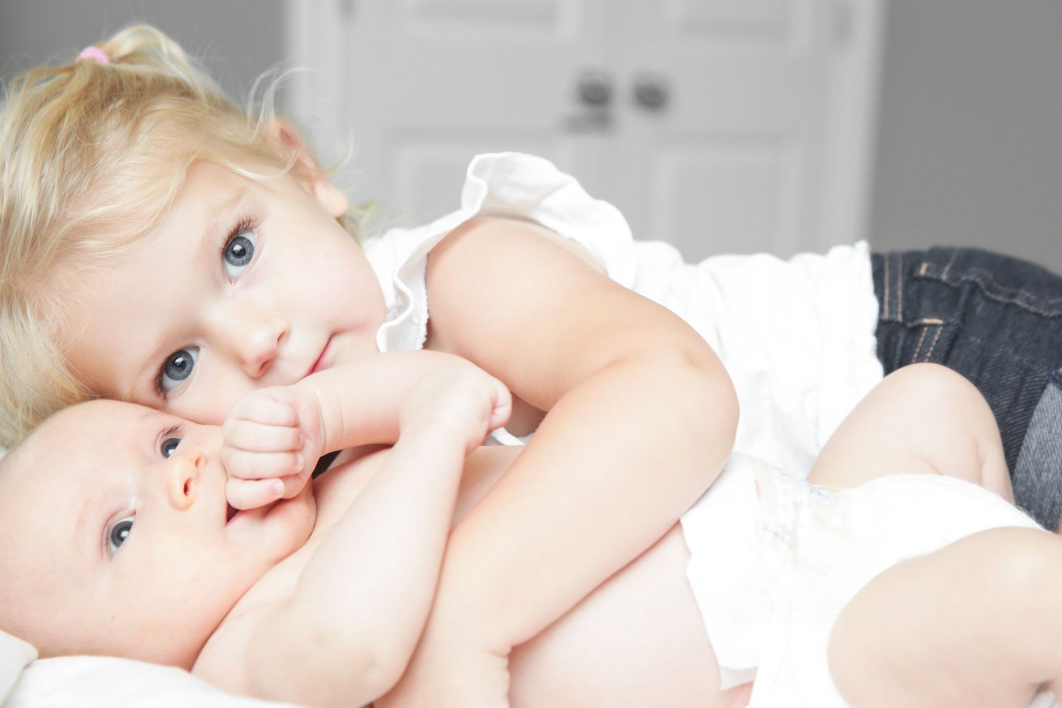 Two blonde toddlers cuddling together, an older child embracing a baby on a white surface.