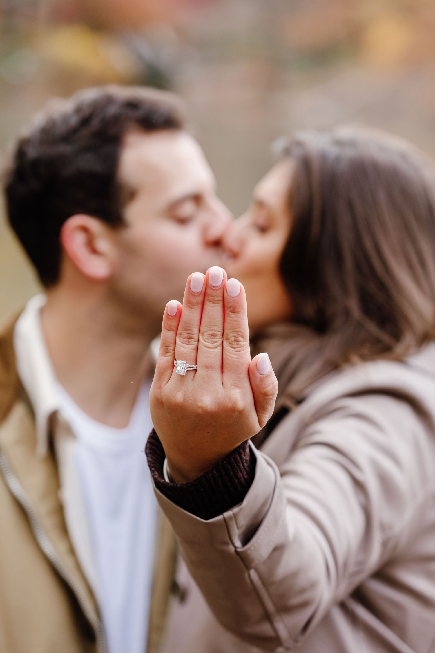 Engaged couple kissing while woman displays diamond engagement ring on her hand in autumn park setting.